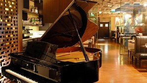 Een vleugelpiano in een ruime, goed verlichte lobby met een houten vloer en een modern plafond.