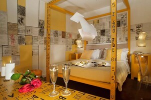 Una camera d'albergo con un letto a baldacchino, un comodino con una lampada, uno sgabello e una parete decorata con vari documenti e schizzi.