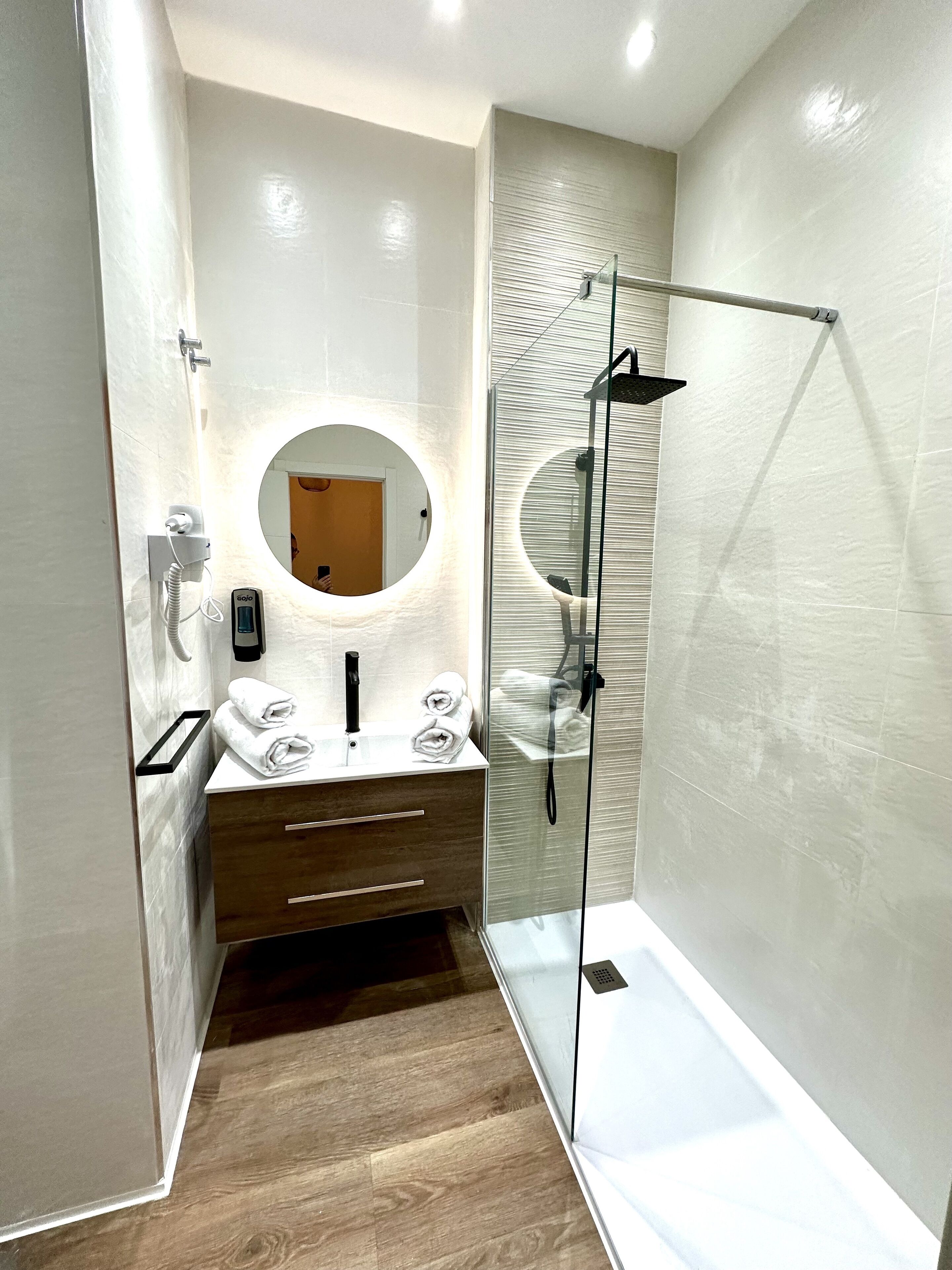 A modern bathroom with a glass-enclosed shower, a round mirror, a wooden vanity, and a towel rack.