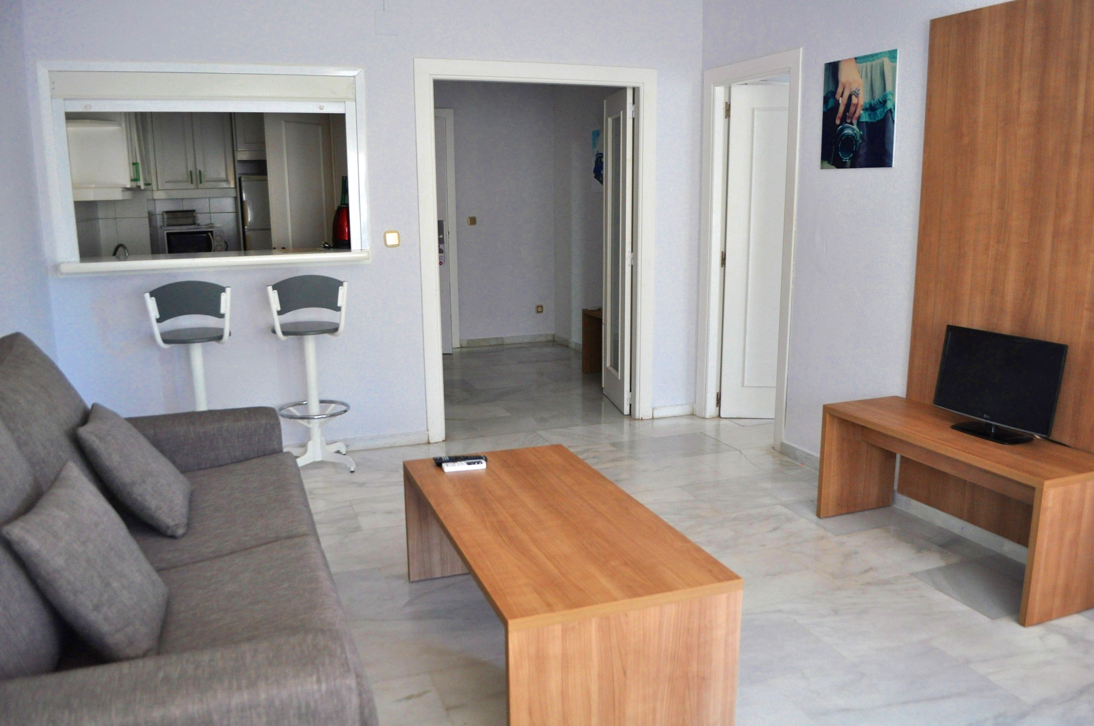 Un salon moderne avec un canapé gris, une table basse en bois, un téléviseur posé sur un meuble en bois, et une cuisine visible à travers une porte ouverte.
