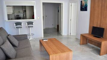 Un salon moderne avec un canapé gris, une table basse en bois, un téléviseur posé sur un meuble en bois, et une cuisine visible à travers une porte ouverte.
