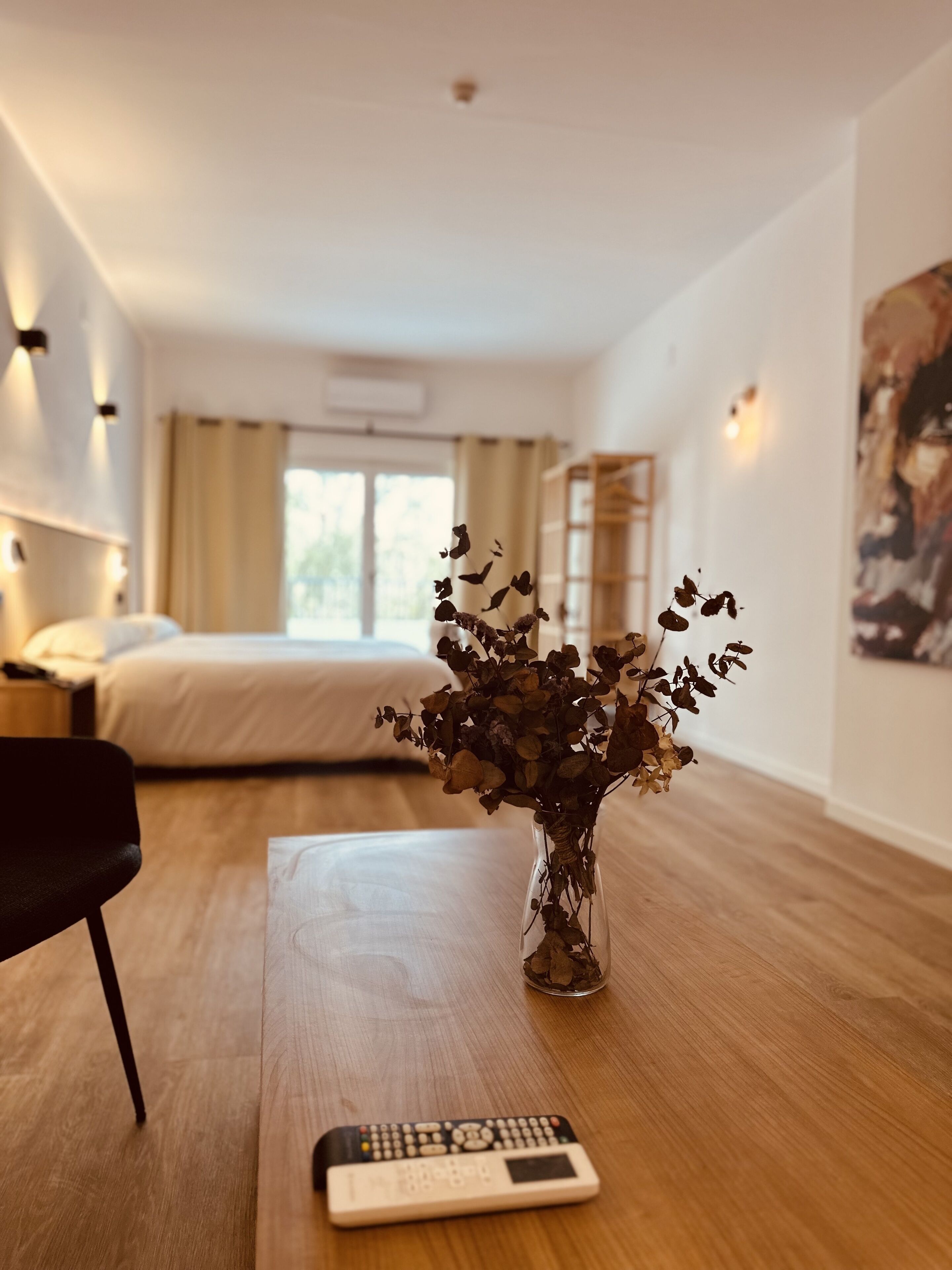 A hotel room with a wooden floor, a bed, a chair, a vase with dried flowers, and a remote control on the table.