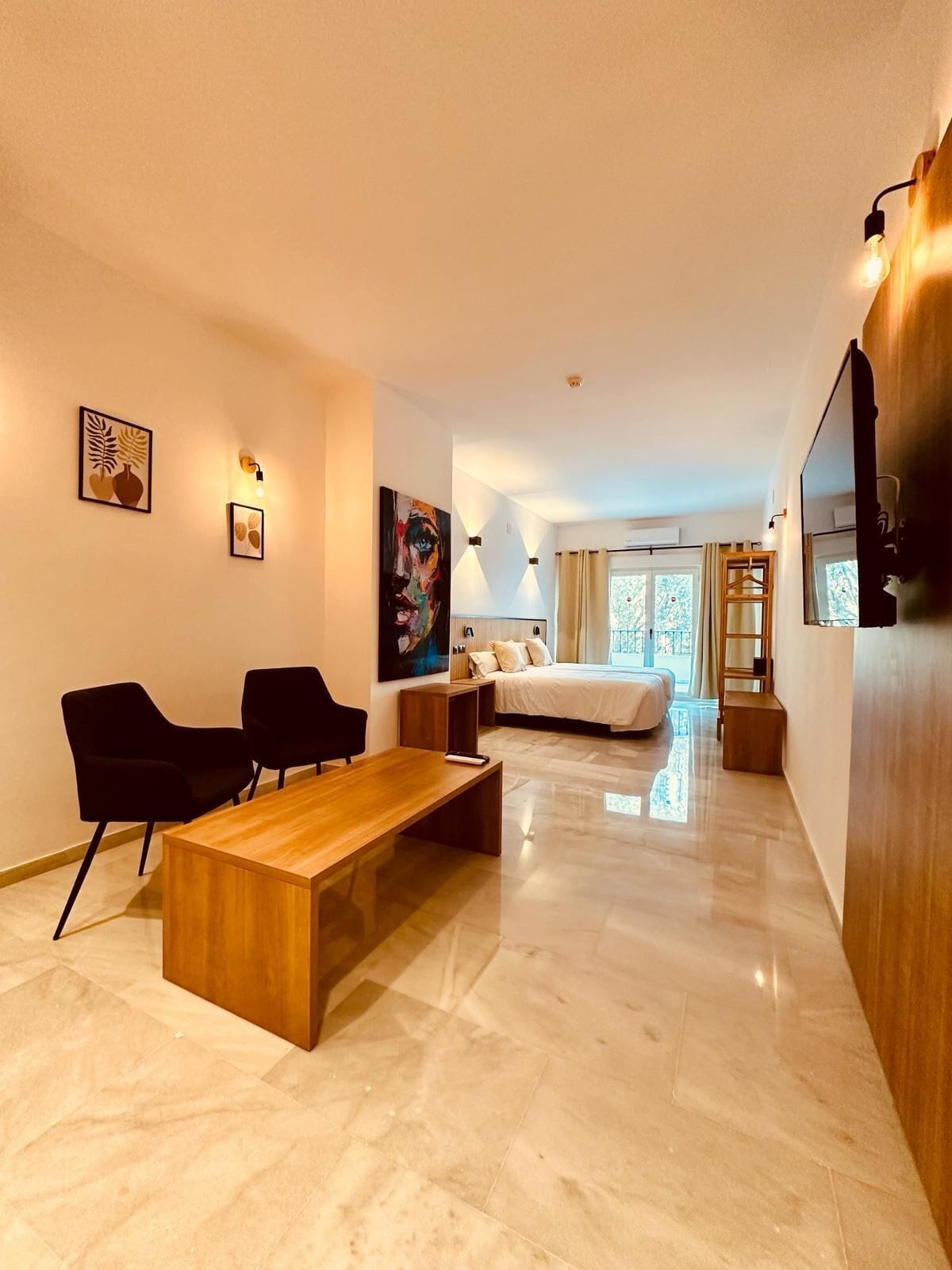A modern hotel room with a wooden coffee table, two black chairs, a bed with white linens, and artwork on the walls.
