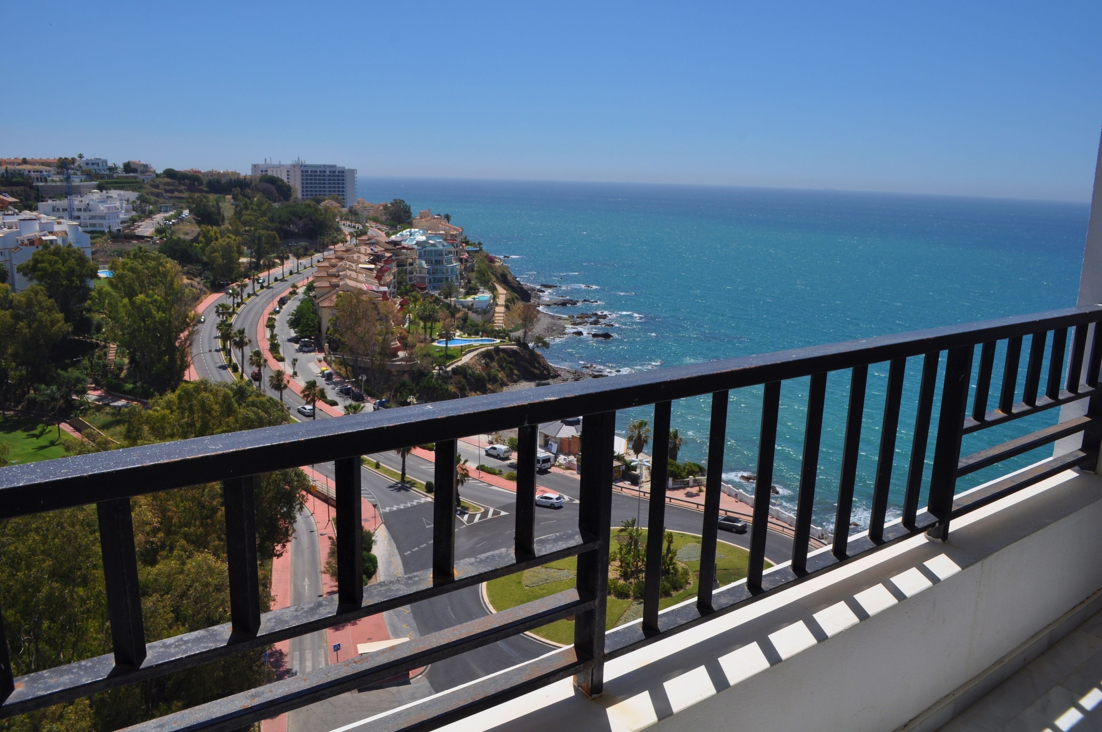 Une vue sur la côte, prise depuis un balcon doté d’une balustrade, donnant sur une route où circulent des véhicules, une piscine et des bâtiments le long de la côte.