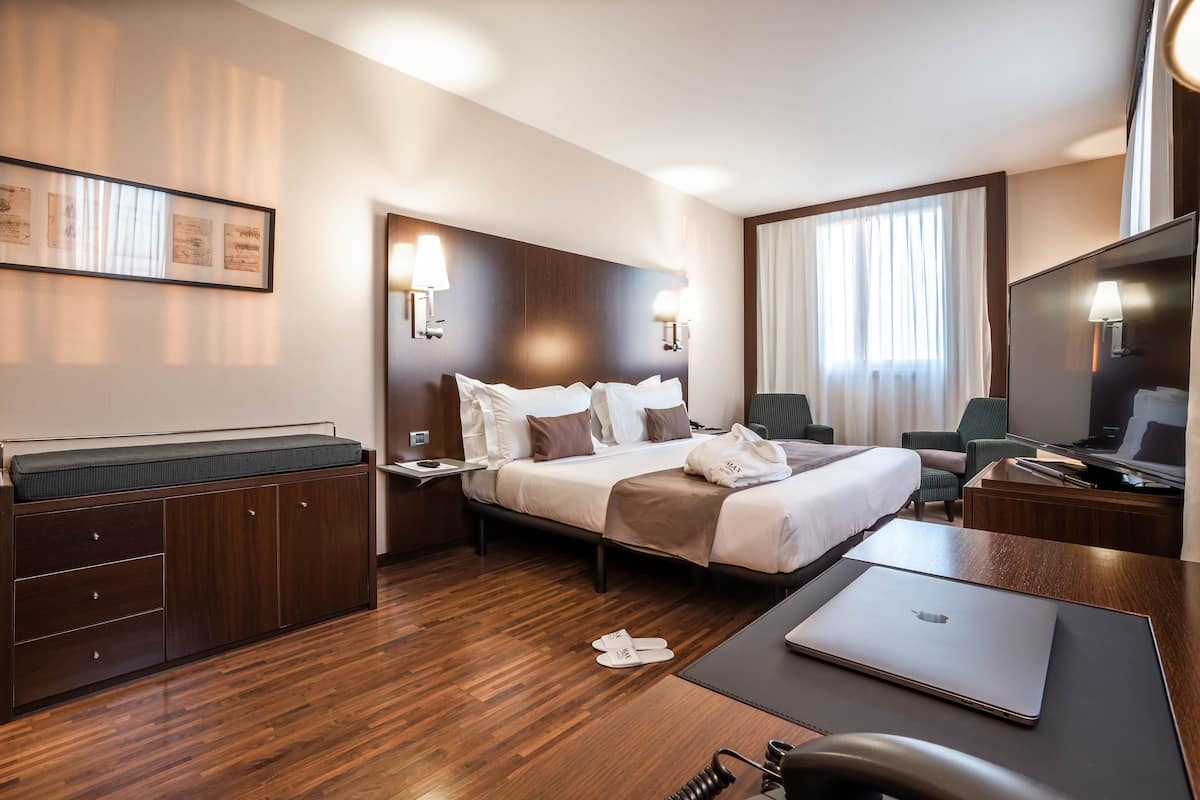 Ein Hotelzimmer mit Bett, Nachttischen, einem Schreibtisch mit Laptop, einem Sessel und einem Fernseher.
