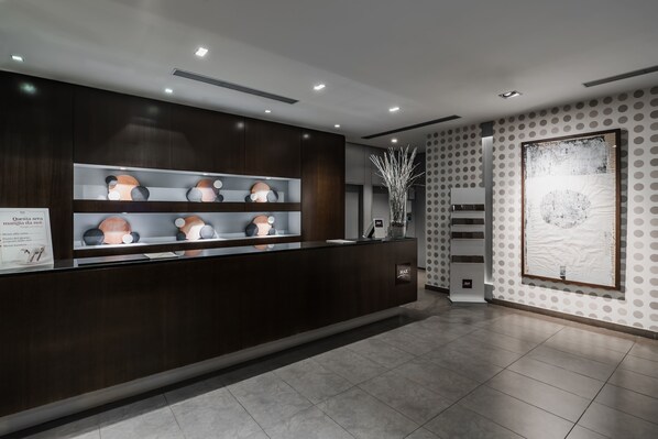 A reception area with a dark wooden counter, shelves with decorative items, and a large framed artwork on the wall. - Max Hotel Livorno (Livorno)