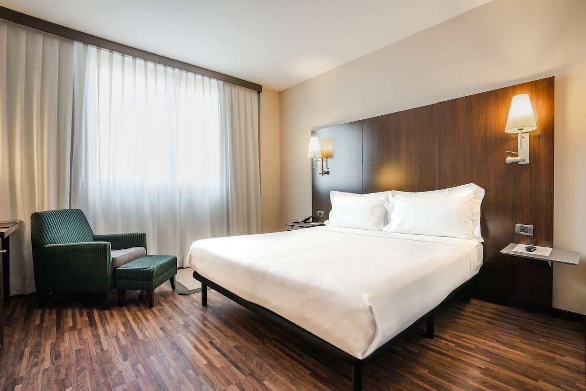 Ein Hotelzimmer mit einem großen Bett, einem grünen Sessel, einem Schreibtisch und einem Nachttisch mit Lampen.