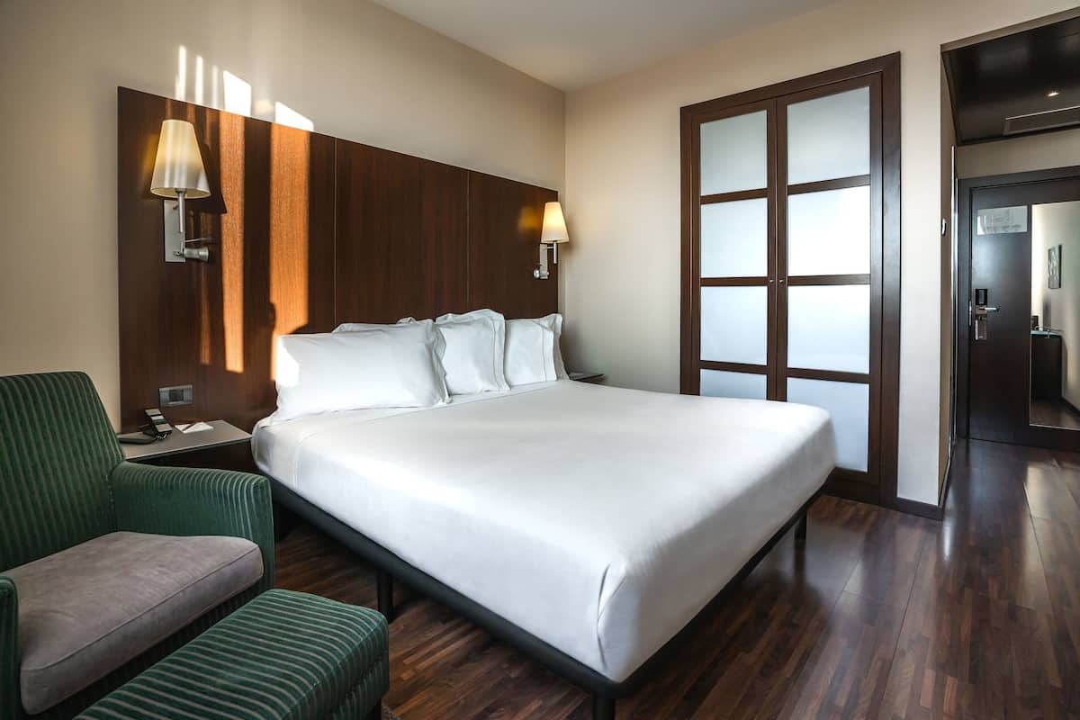 Ein Hotelzimmer mit einem großen Bett, einem grünen Sessel, einem Nachttisch und einer Wandlampe.