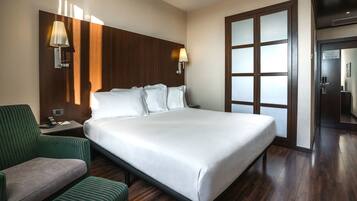 A hotel room with a large bed, a green armchair, a nightstand, and a wall-mounted lamp.