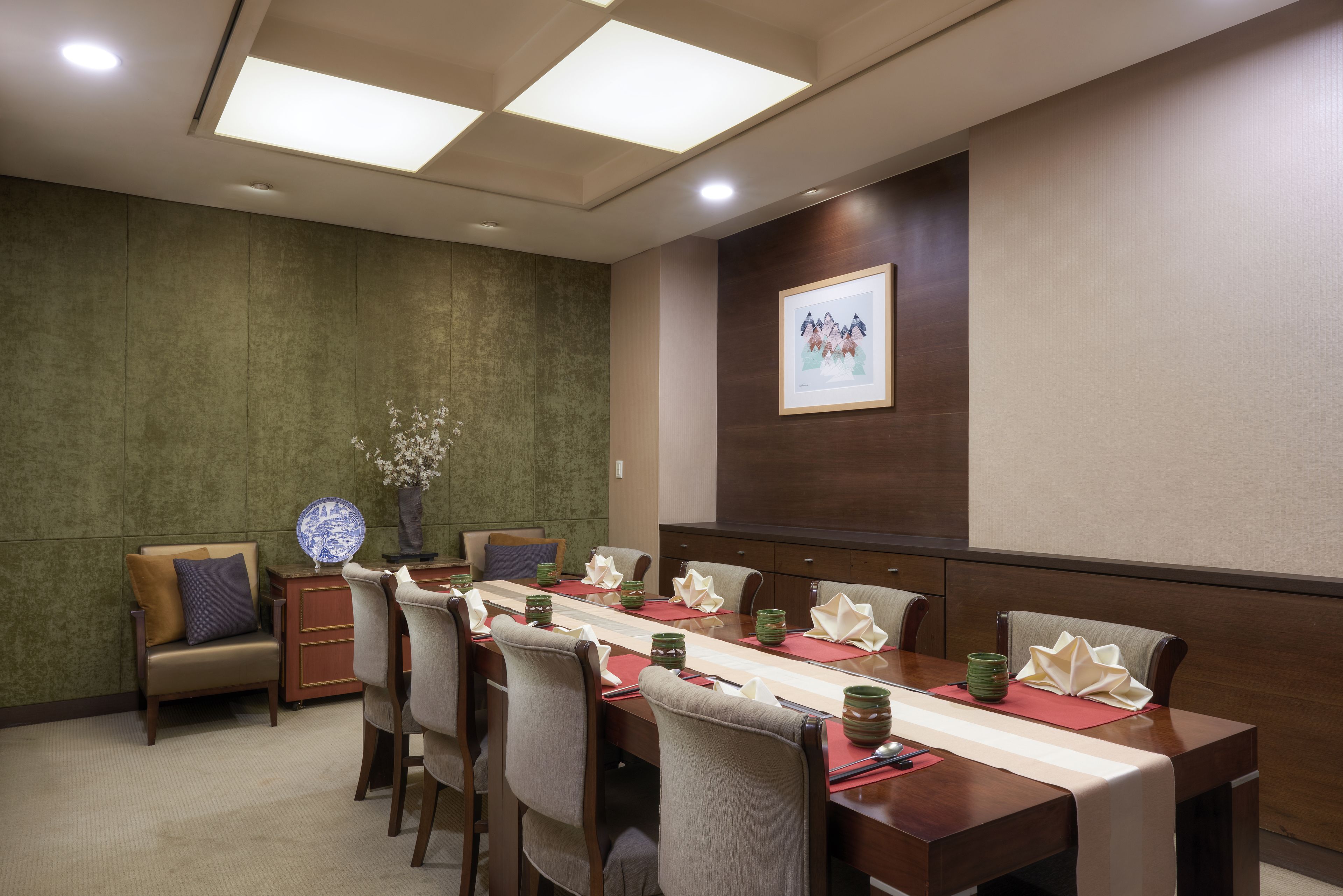 Un salón de conferencias con una larga mesa, sillas, un florero con flores y un cuadro en la pared.
