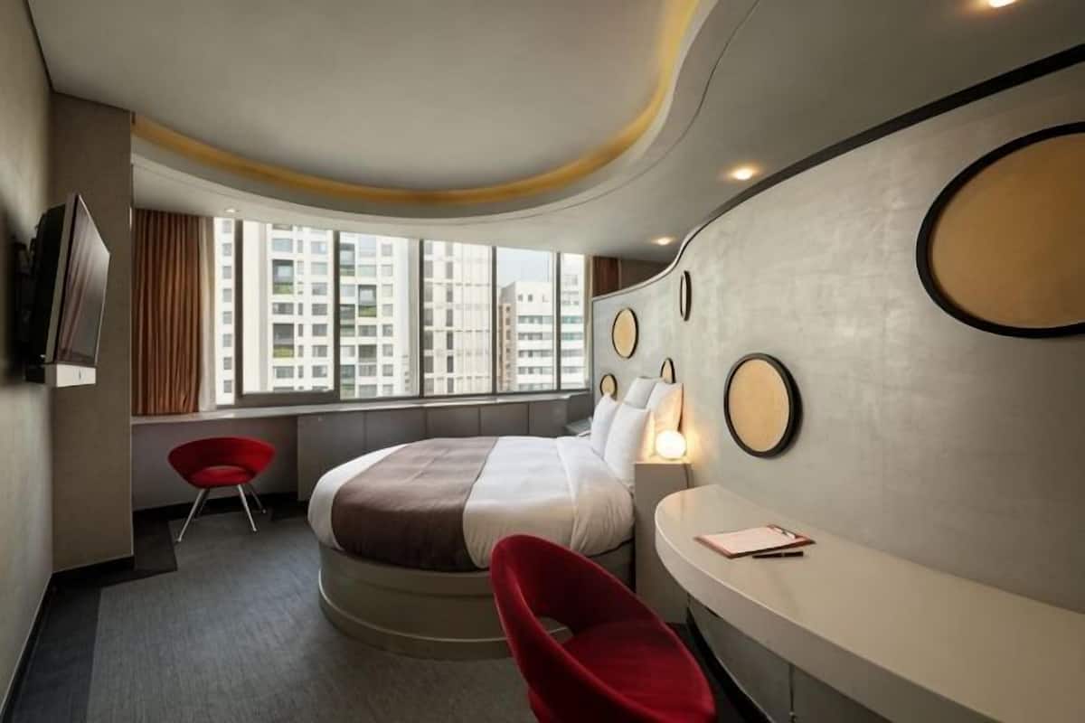 A modern hotel room with a curved ceiling, a bed, a desk, a chair, and a large window with a city view.