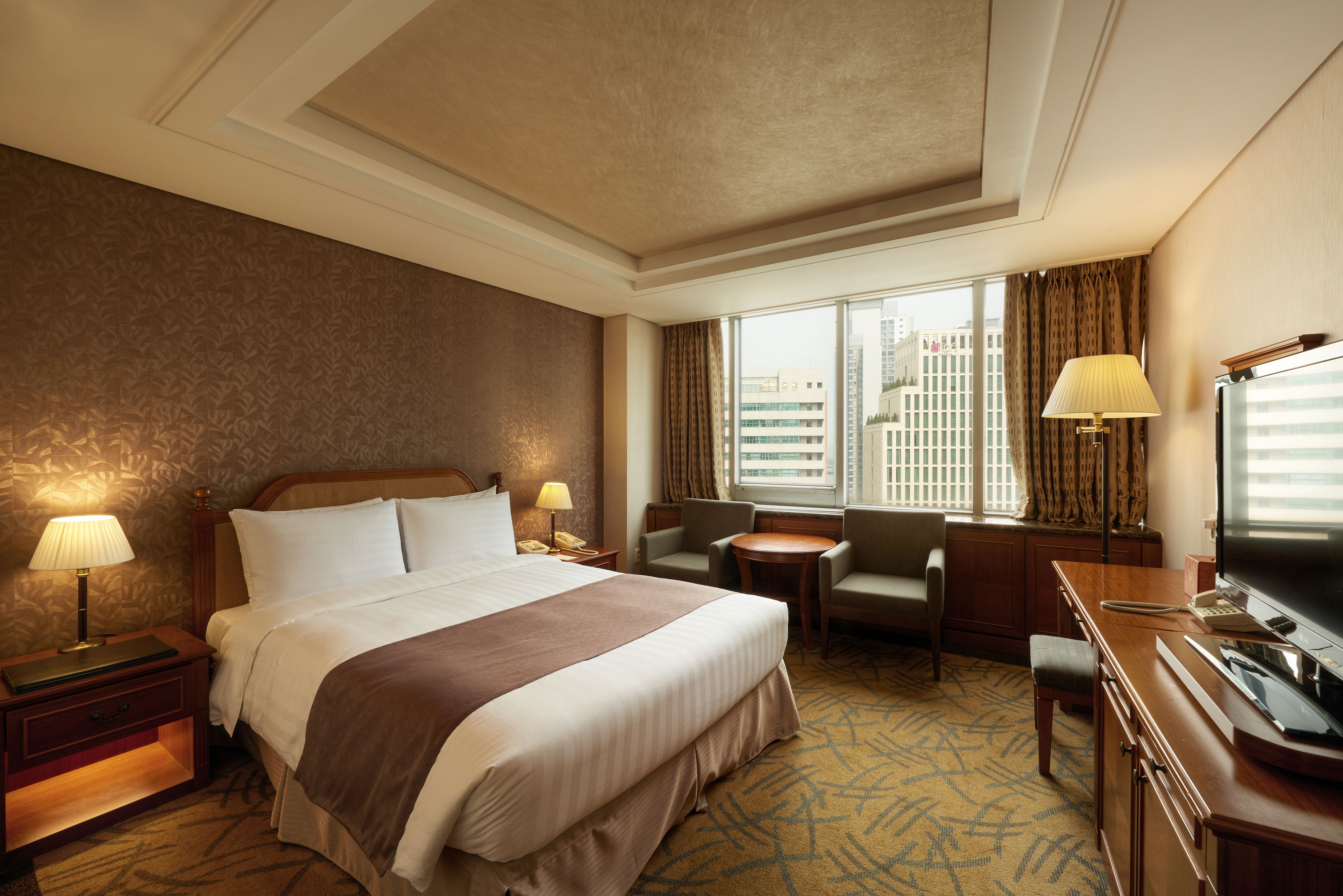 Une chambre d’hôtel avec un grand lit, deux tables de chevet avec des lampes, un bureau avec une télévision, une chaise et une vue sur les bâtiments par la fenêtre.