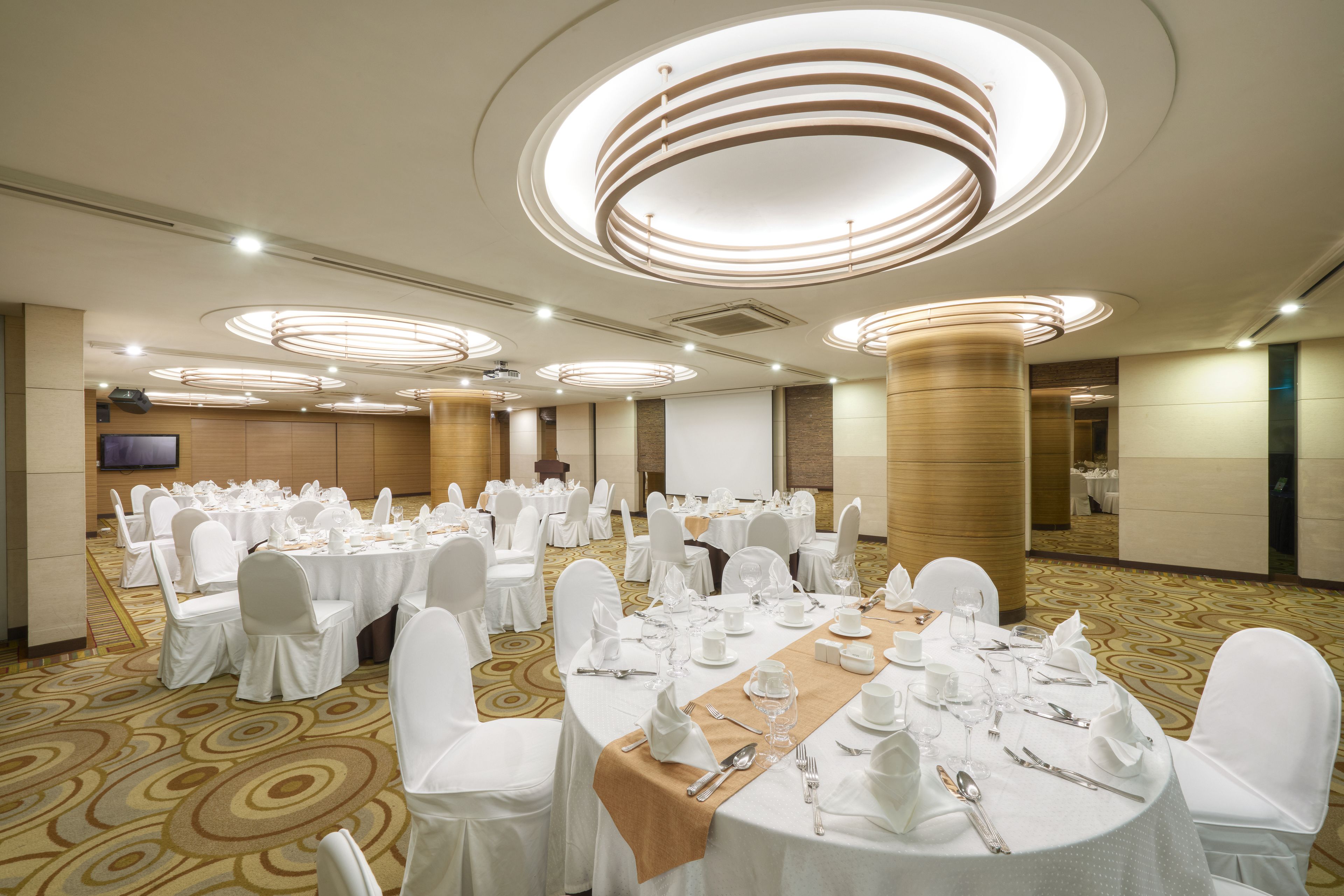 A banquet hall with round tables set for a formal event, featuring a central stage and a large screen.