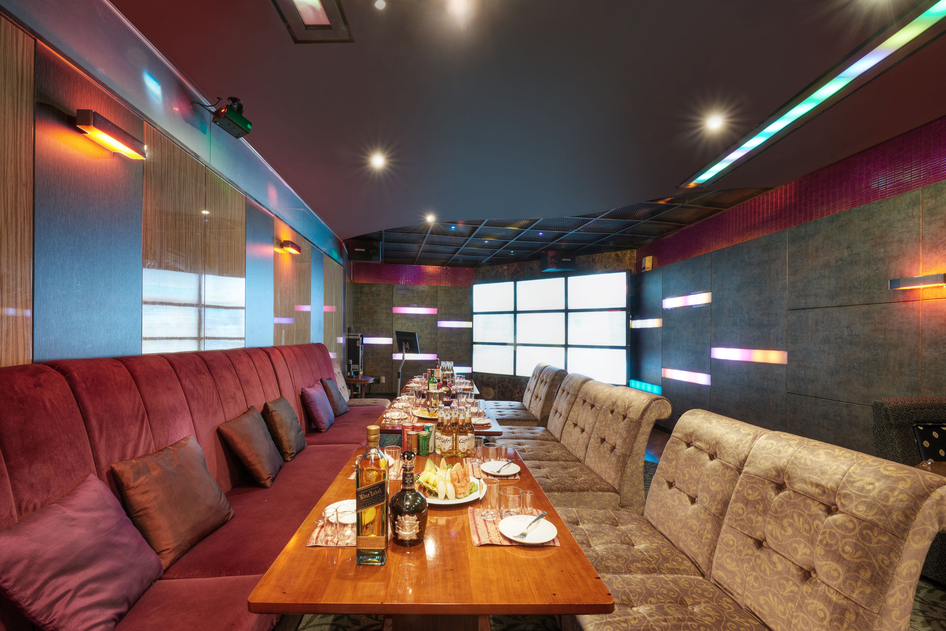 Una larga mesa de comedor dispuesta con platos, vasos y botellas, flanqueada por sofás tapizados en rojo y beige.