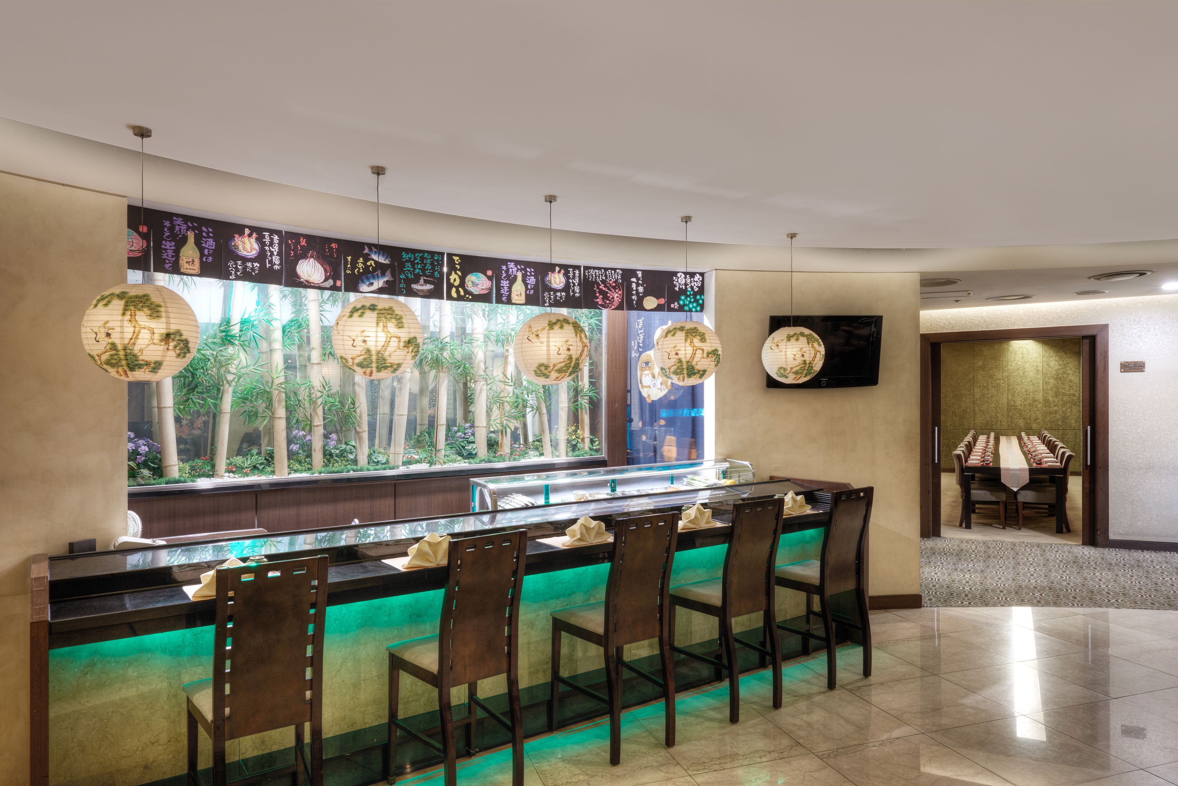A restaurant with a sushi bar, green ambient lighting, and a view of bamboo plants.