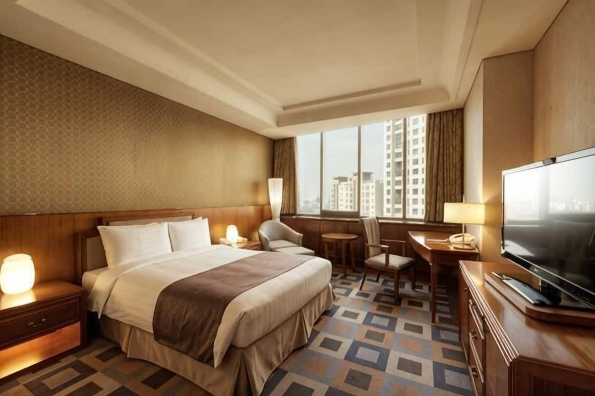 A hotel room with a large bed, bedside tables, a desk, a chair, a television, and a window with a city view.