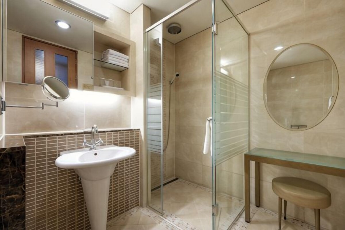 Een badkamer met een glazen douchecabine, een wastafel op een zuil, een ronde spiegel en een wand betegeld met marmer.