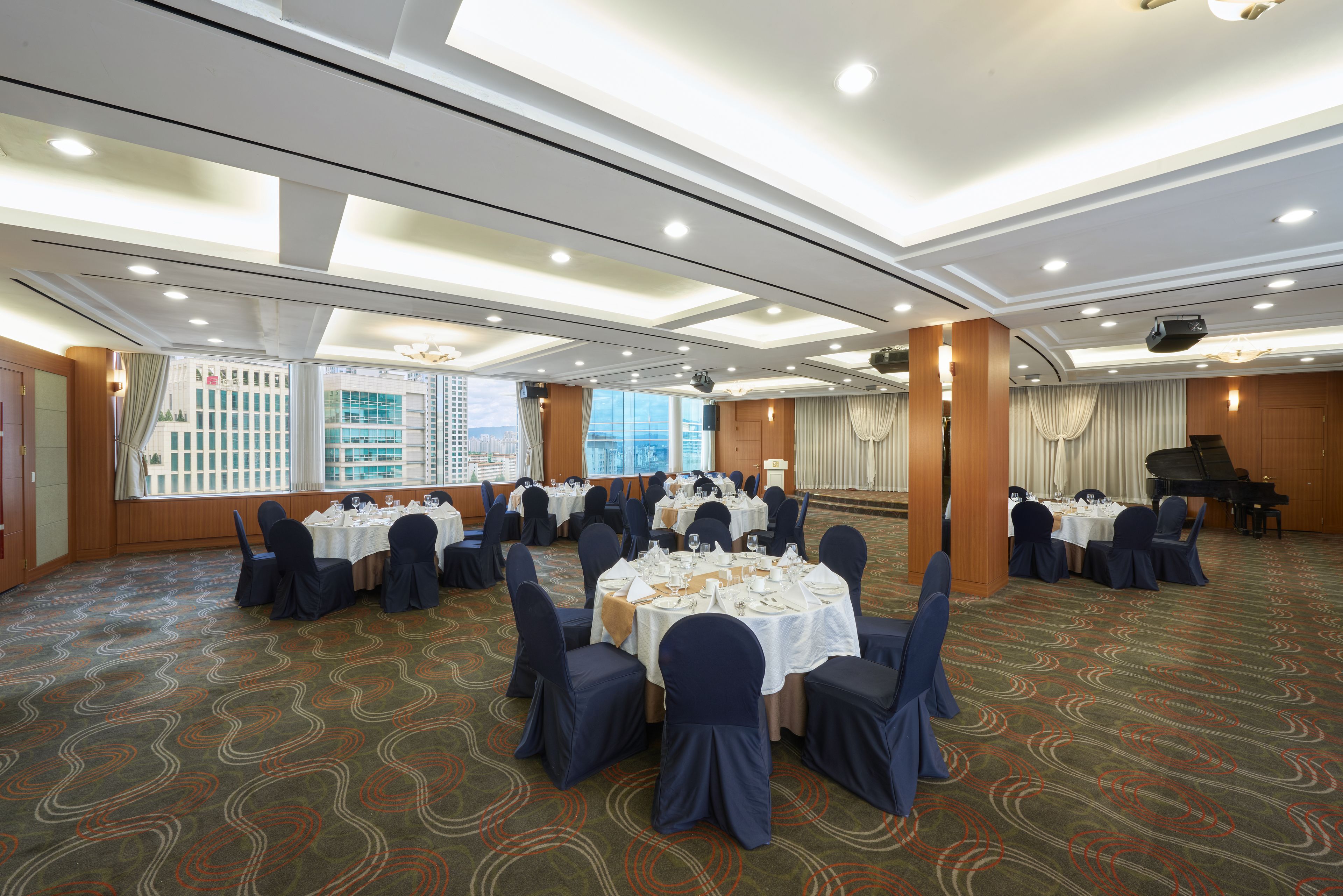 A banquet hall with round tables set for dining, a stage with a piano, and large windows offering a city view.