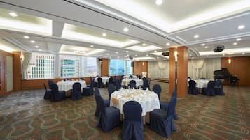 A banquet hall with round tables set for dining, a stage with a piano, and large windows offering a city view.