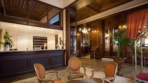 A reception area with a wooden reception desk, several upholstered chairs, a chandelier, and a large mirror. - Grand Hotel Moderne (Lourdes)