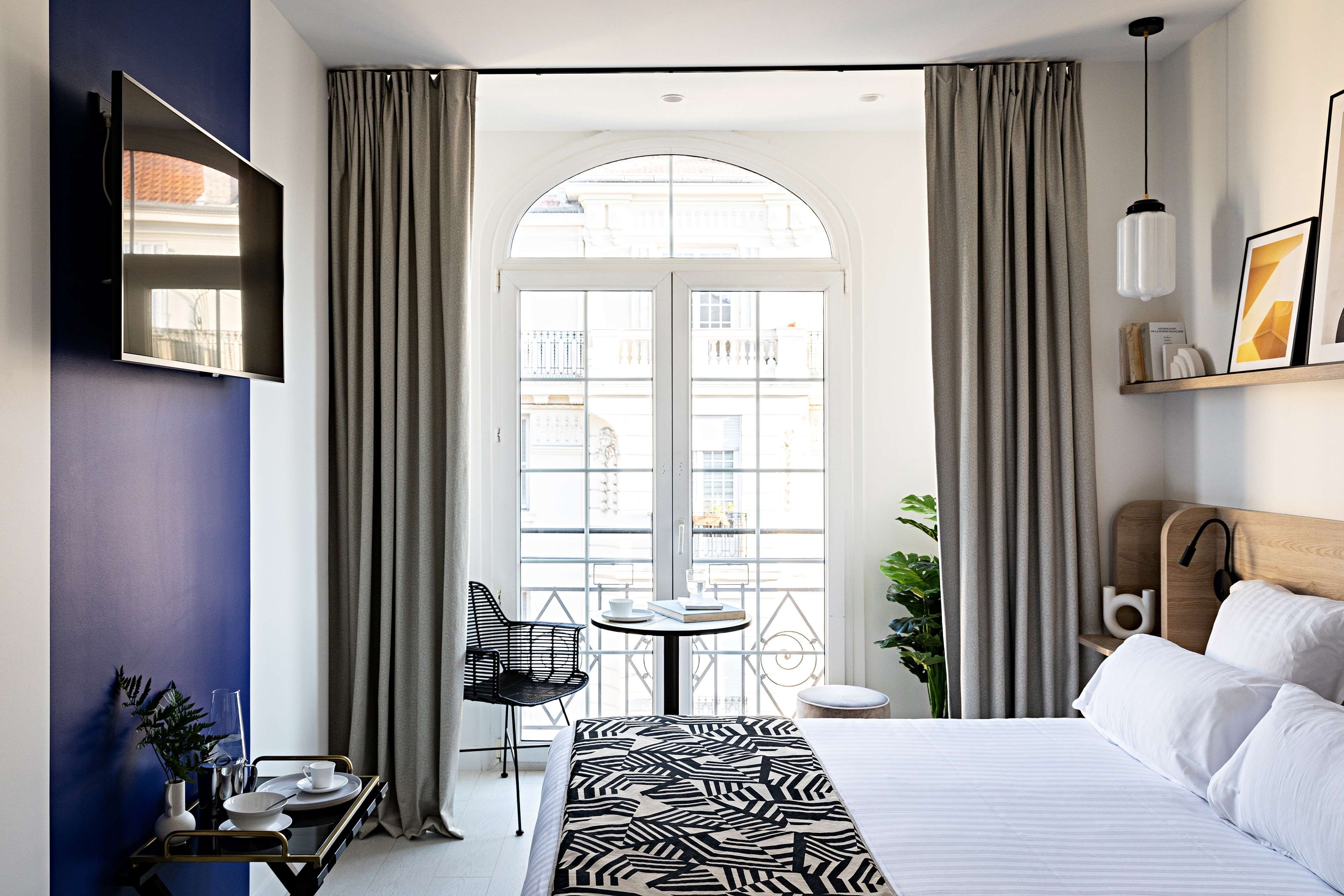 A hotel room with a bed, a desk with a chair, a small table with a chair, a plant, and a view through a window.