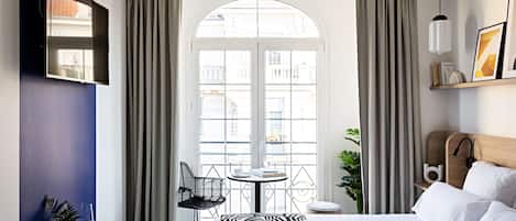 A hotel room with a bed, a desk with a chair, a small table with a chair, a plant, and a view through a window.