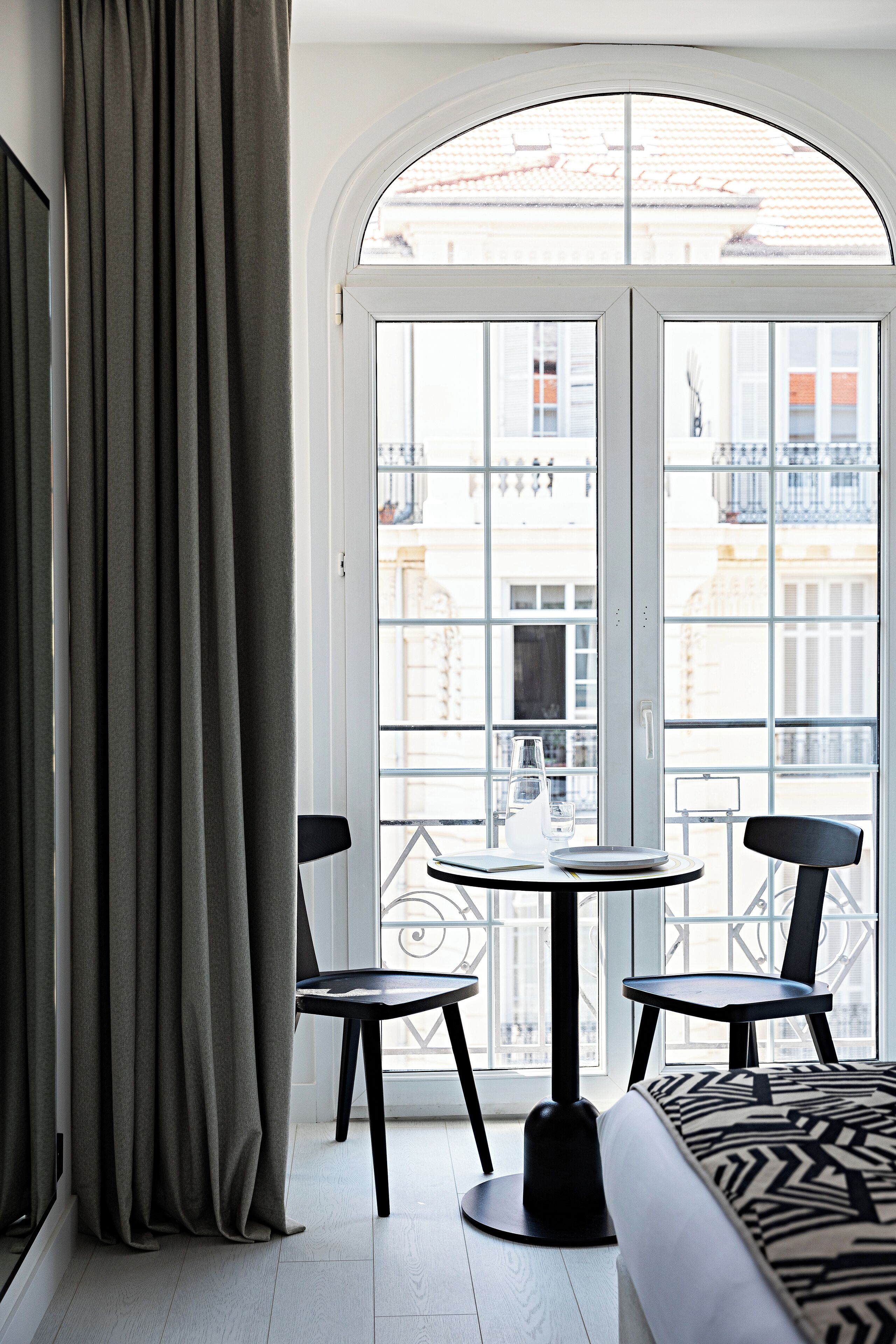 Una habitación con cama, una mesa y sillas, un ventanal con cortinas y vistas a edificios.