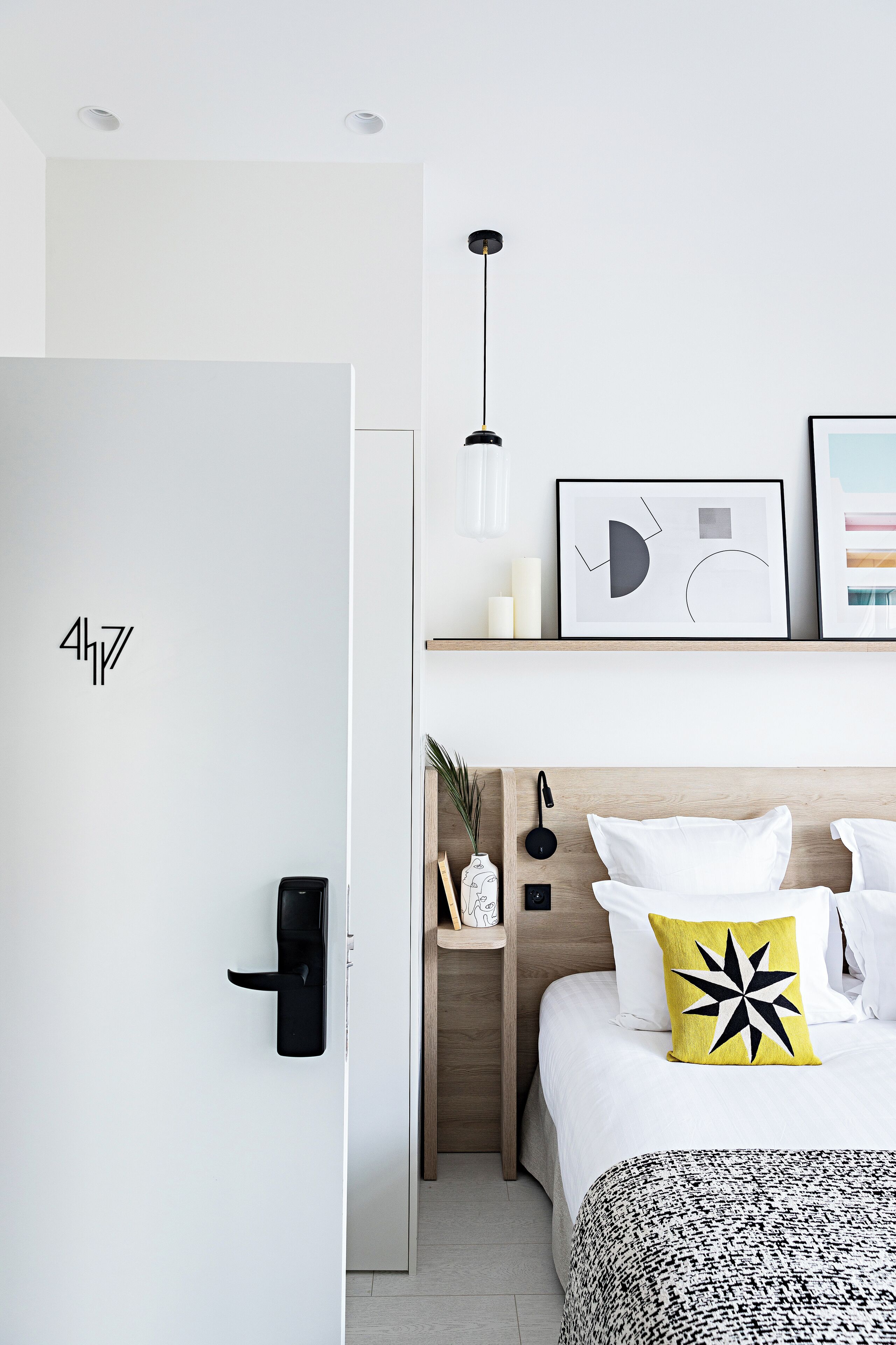 A modern hotel room with a minimalist design, featuring a bed with a patterned bedspread, a bedside table with a lamp, and a wall-mounted shelf with framed artwork and a vase.