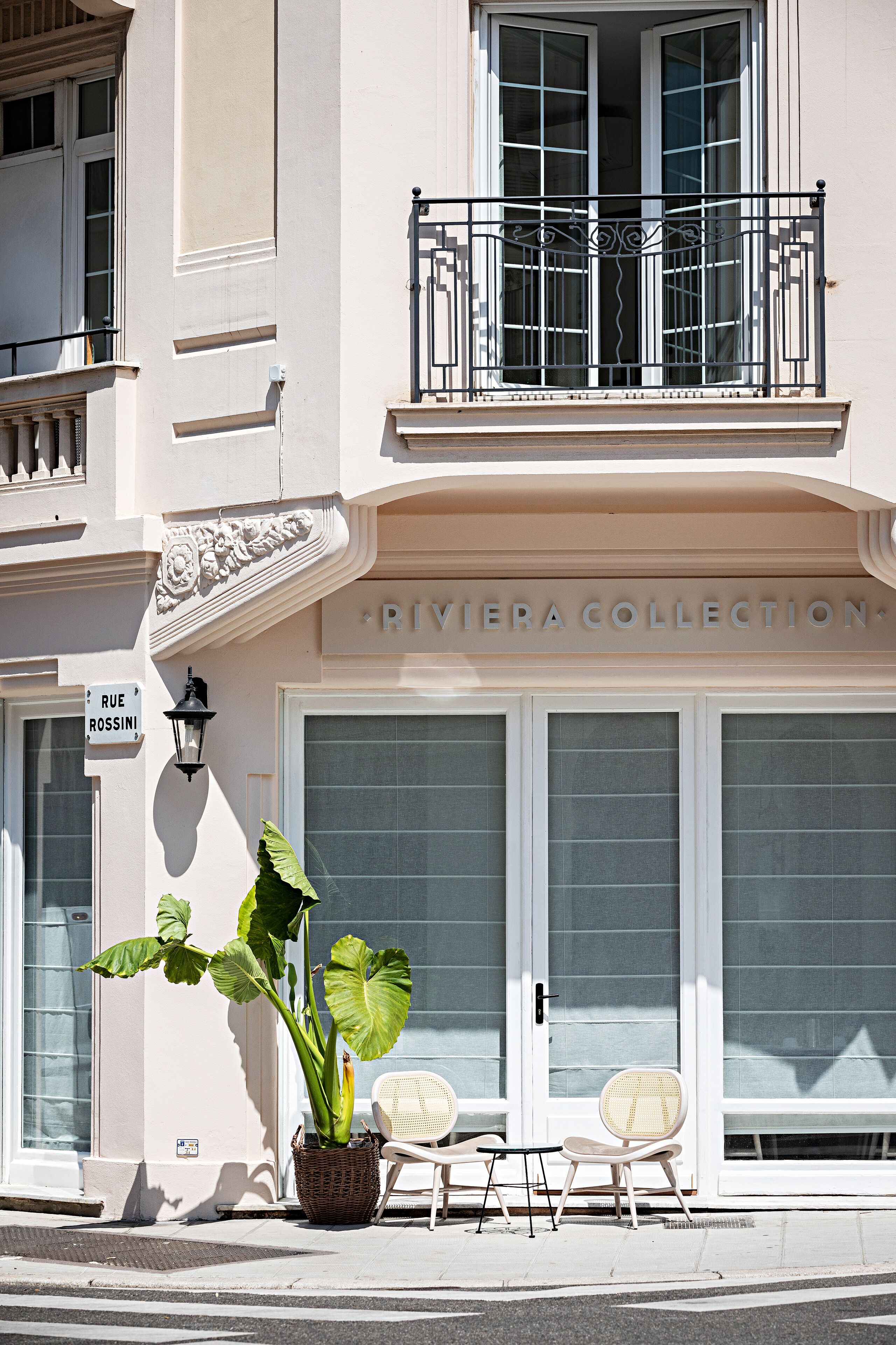 A building with a sign reading "RIVIERA COLLECTION" and a sign for "RUE ROSSINI". There is a potted plant, two wicker chairs, and a small table outside the building.