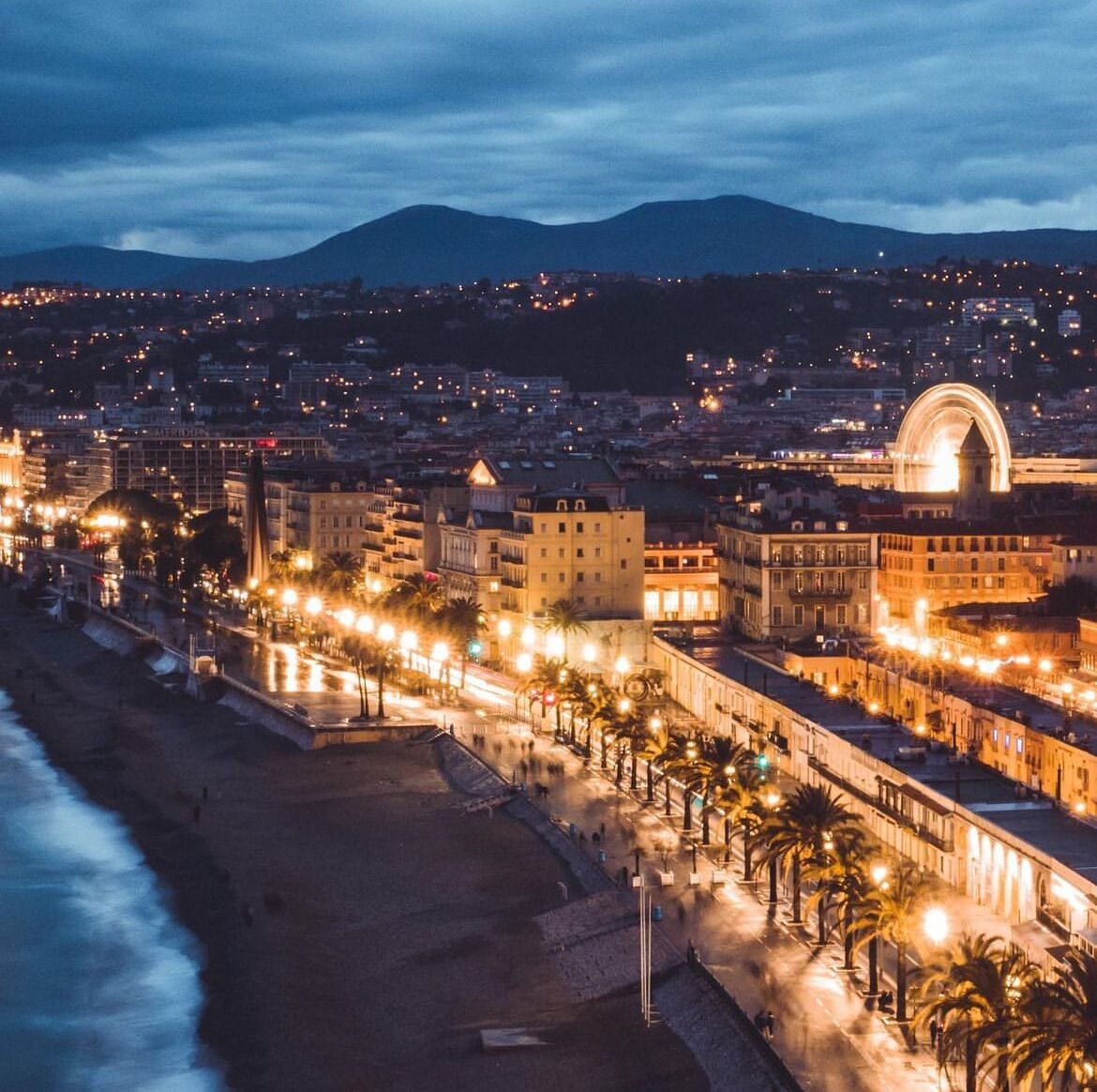 Paisagem costeira ao entardecer com uma estrutura em arco imponente, uma praia e um calçadão bem iluminado.