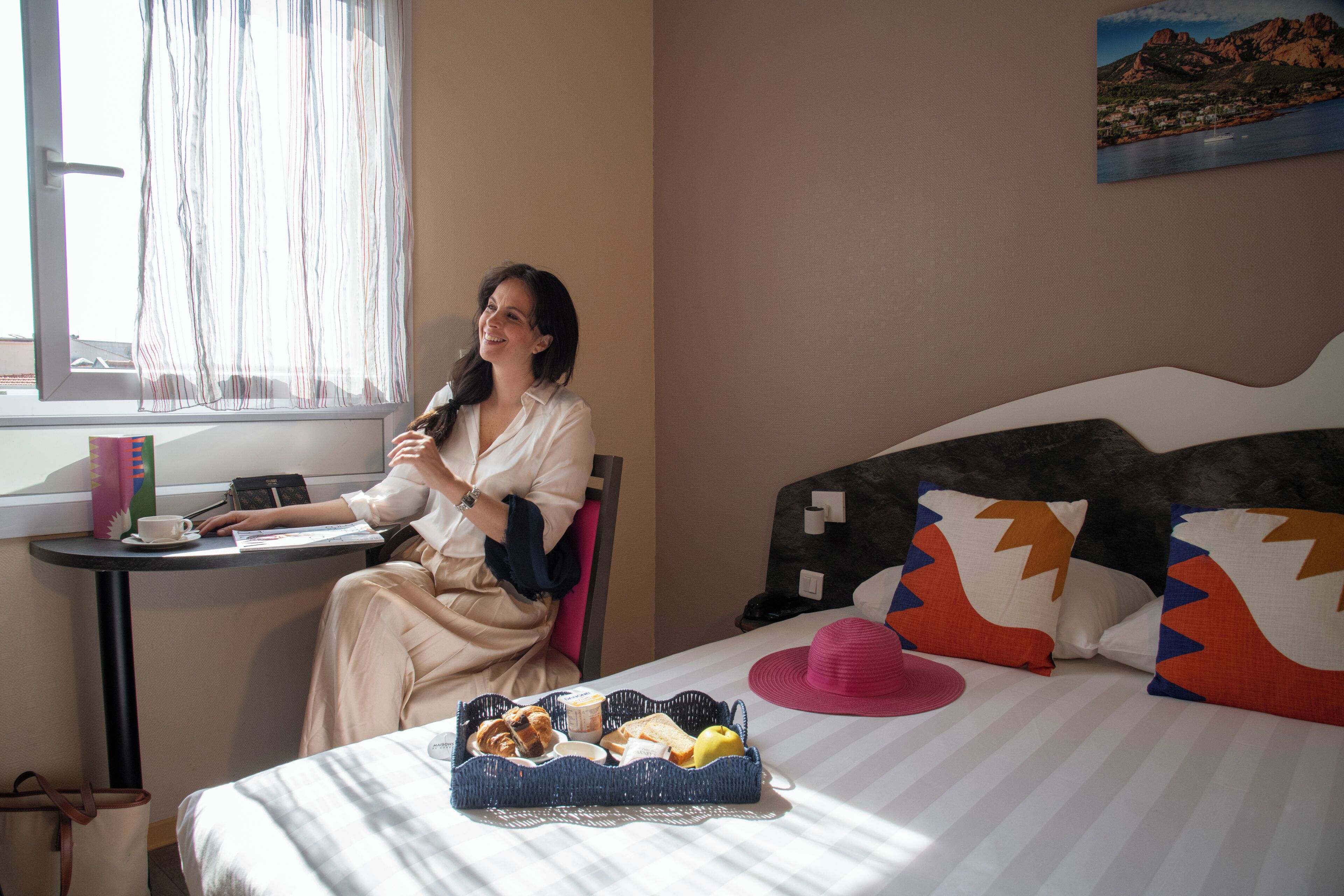 A hotel room with a bed, a table with breakfast items, a window with curtains, and a painting on the wall.