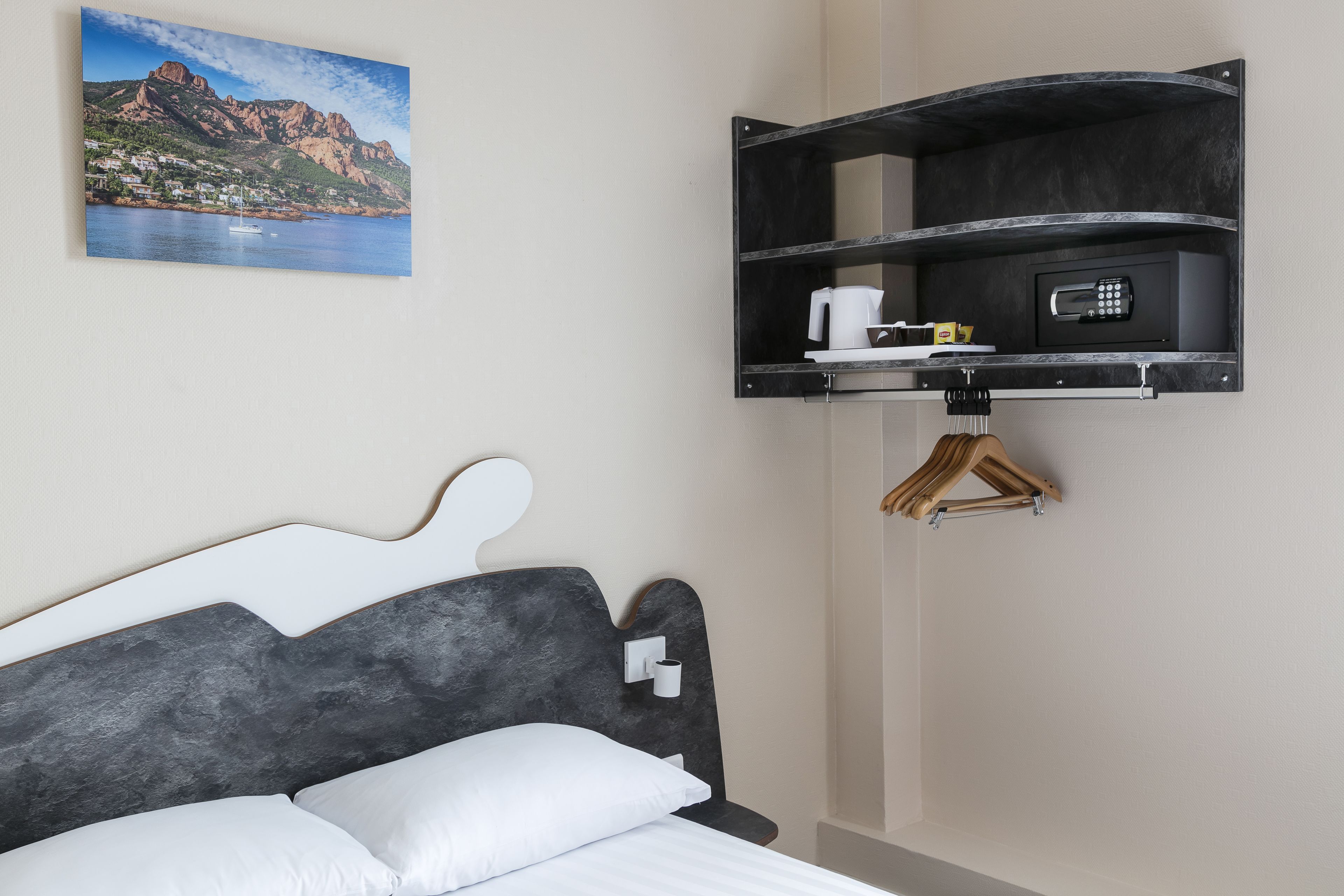 A hotel room with a bed, a mounted shelf with a coffee maker, a remote control, and a wooden hanger.