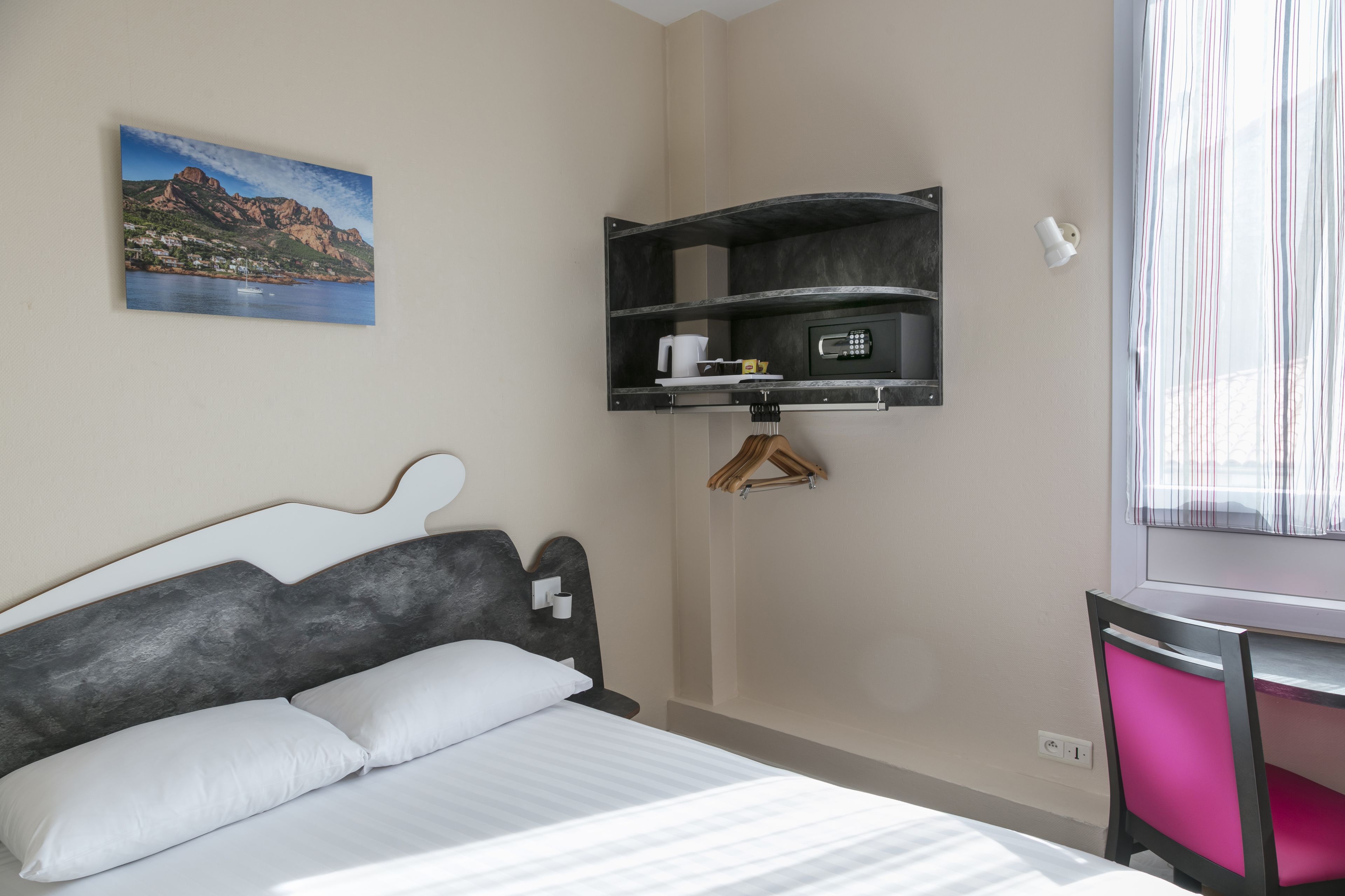 Un dormitorio con cama, escritorio, estante con hervidor y un cuadro de un paisaje montañoso.