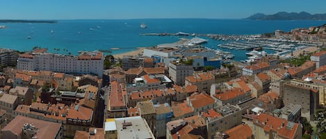 Un paisaje urbano costero con arquitectura densa, un puerto deportivo con numerosos barcos, y una vista despejada del mar y montañas distantes.