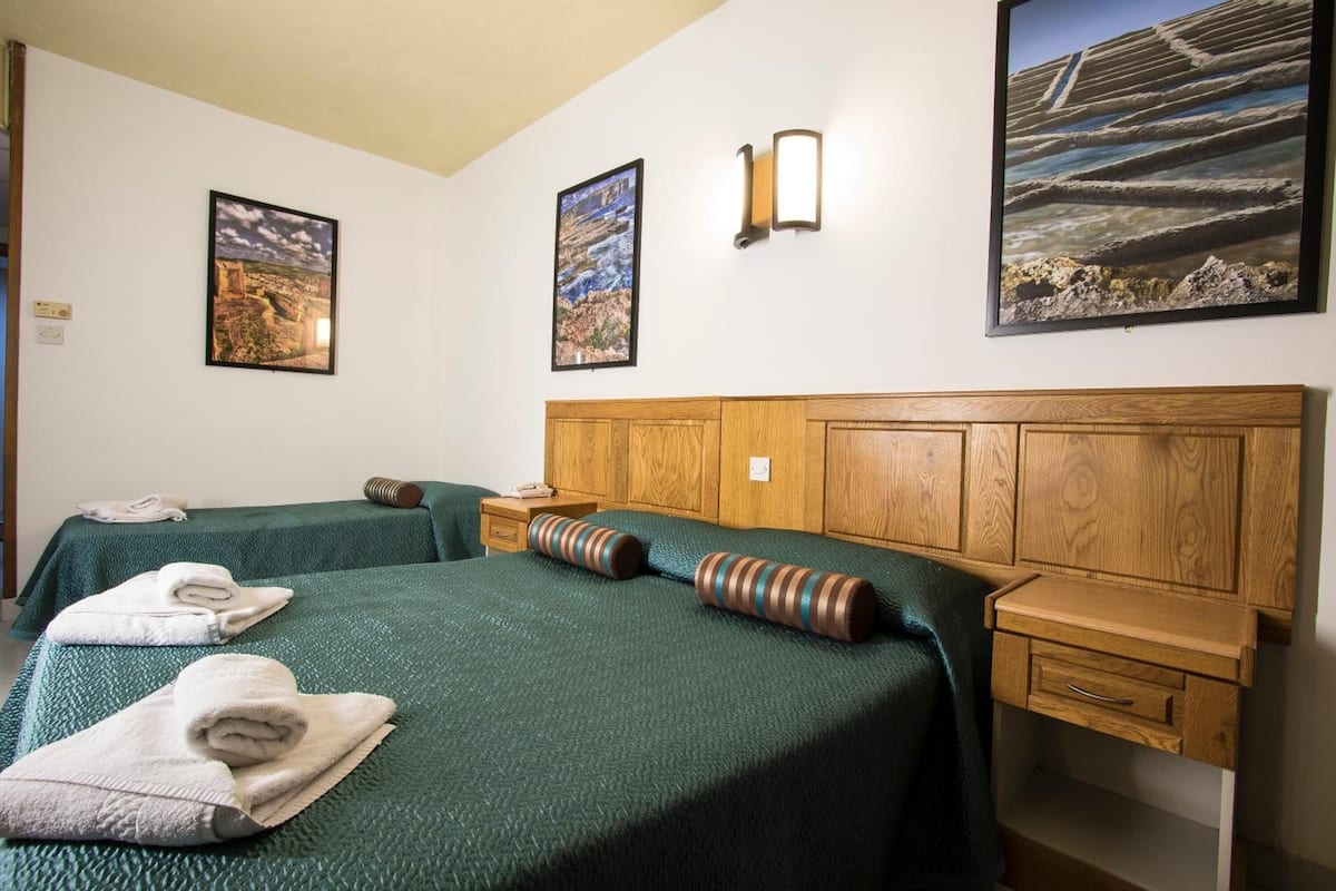 Habitación de hotel con dos camas, cabeceros de madera, una mesita de noche y dos lámparas fijadas a la pared.