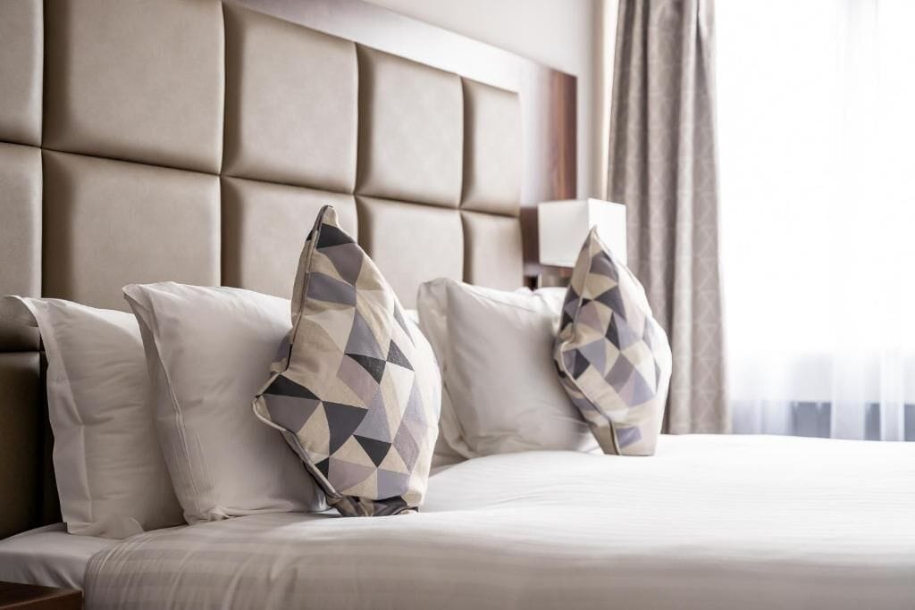 A hotel room with a large padded headboard, a bed with white bedding, and decorative pillows.