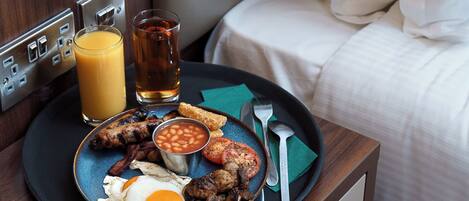 A breakfast tray with eggs, bacon, beans, and toast on a wooden table next to a bed with a white headboard.