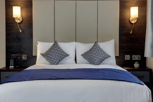 Ett hotellrum med en stor säng, två mönstrade kuddar, ett blått täcke och väggmonterad belysning.