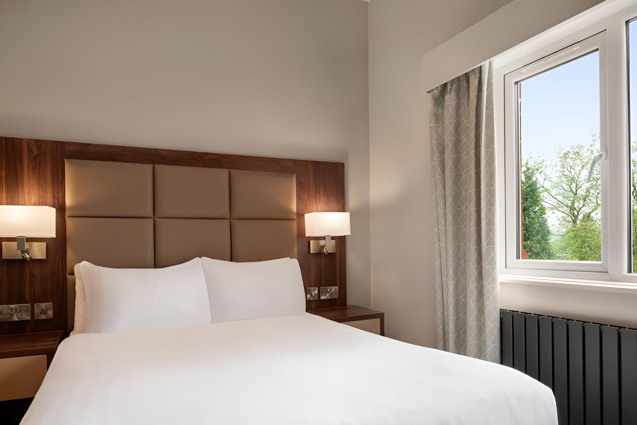 A hotel room with a large bed, two bedside lamps, a window with curtains, and a wooden headboard.