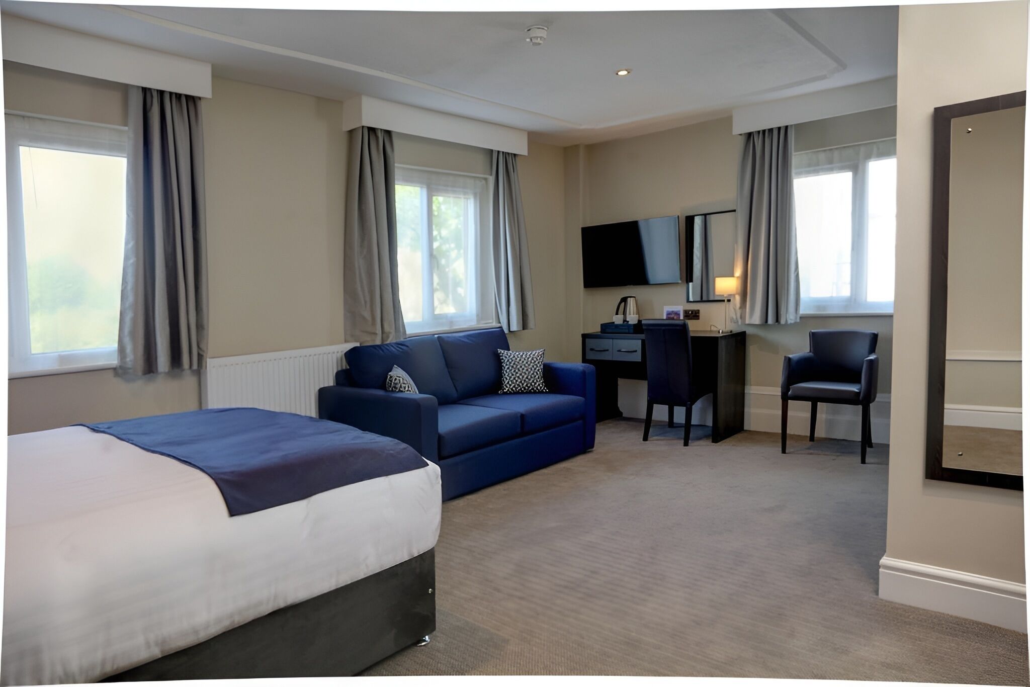 Habitación de hotel con cama, sofá azul, escritorio con silla, televisión y ventanas amplias con cortinas.