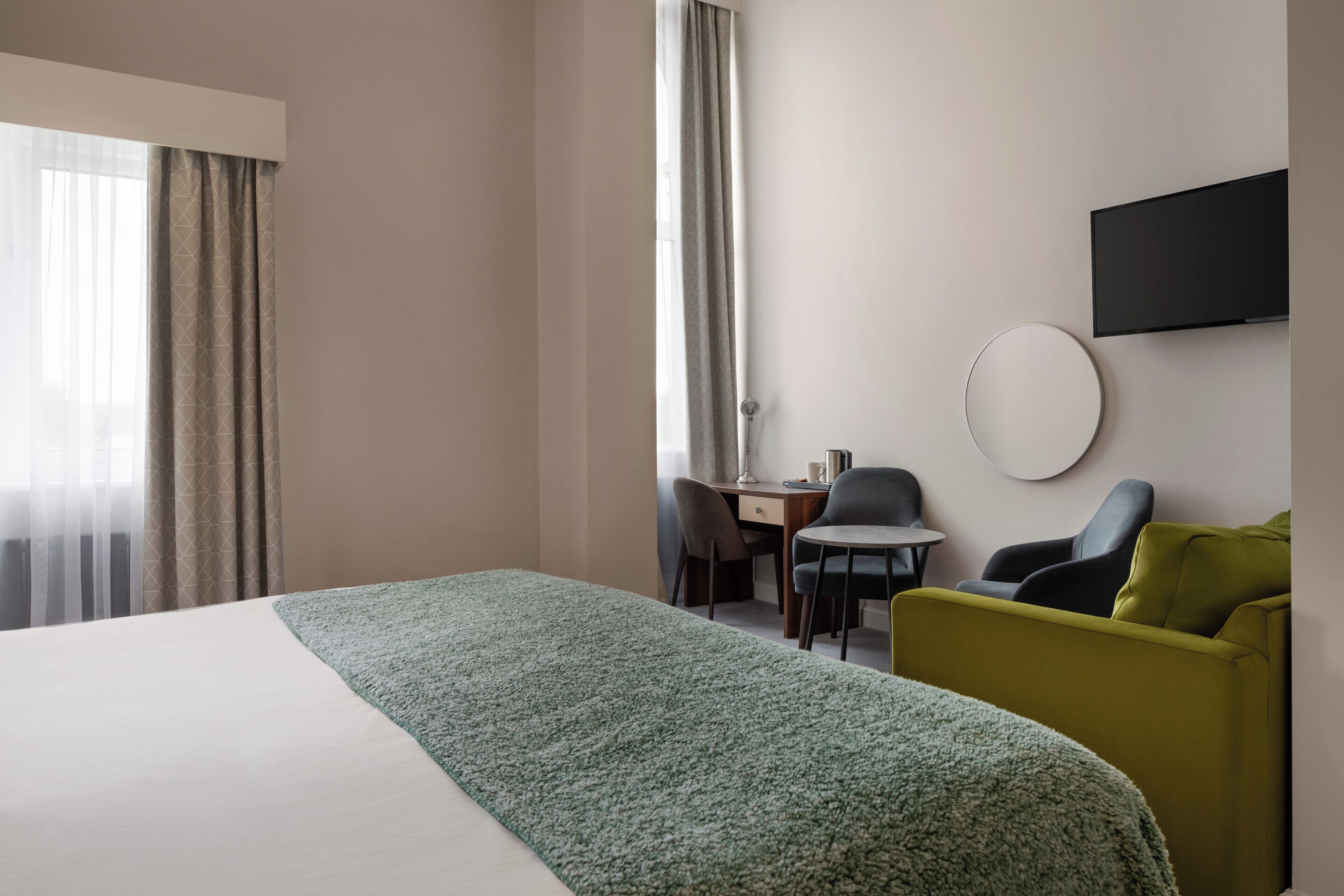 A hotel room with a bed, a desk, two chairs, a TV, and a window with curtains.