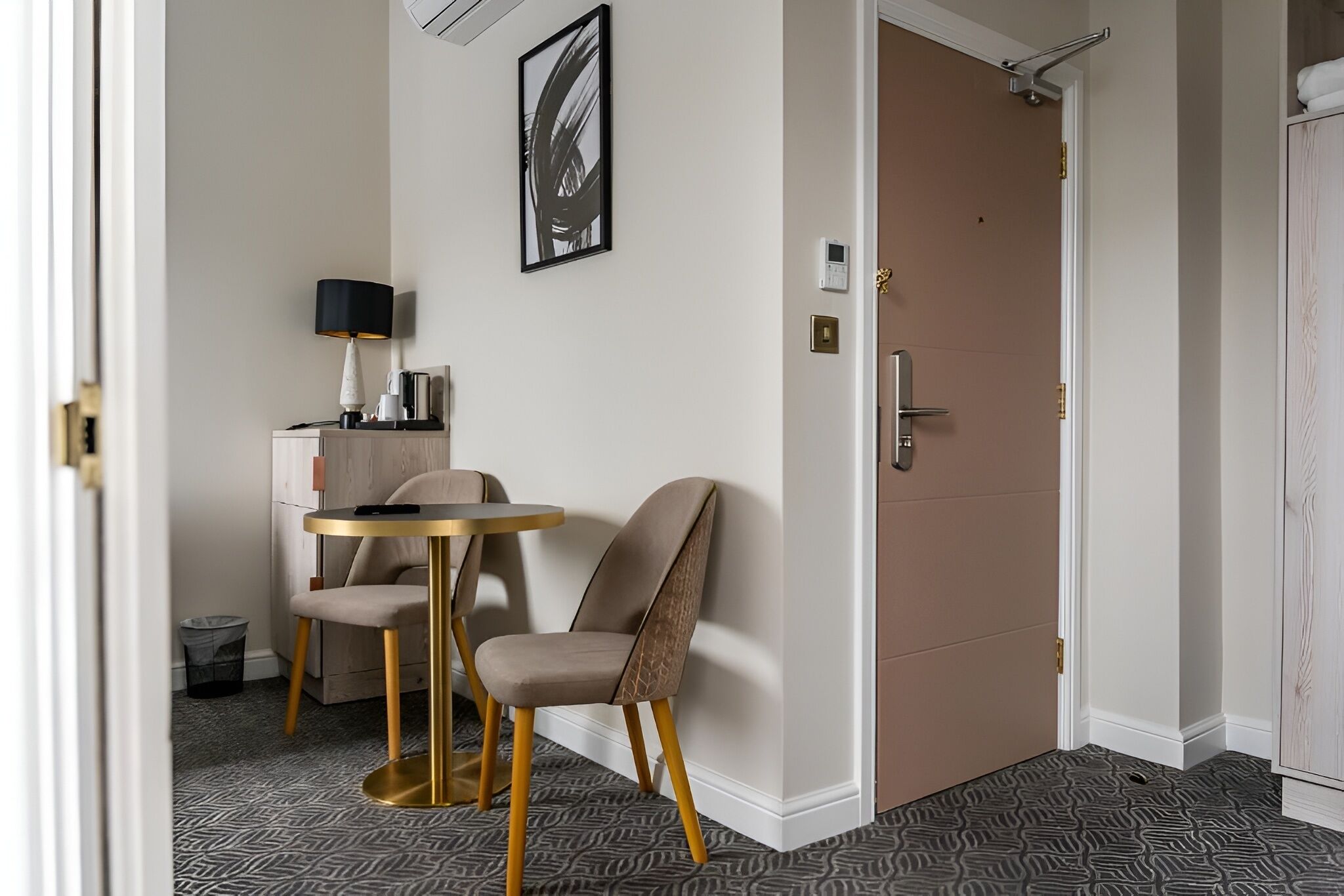 Ein Hotelzimmer mit rundem Tisch, zwei Stühlen, einer Lampe, einem Bild, einer Tür und einem gemusterten Teppich.