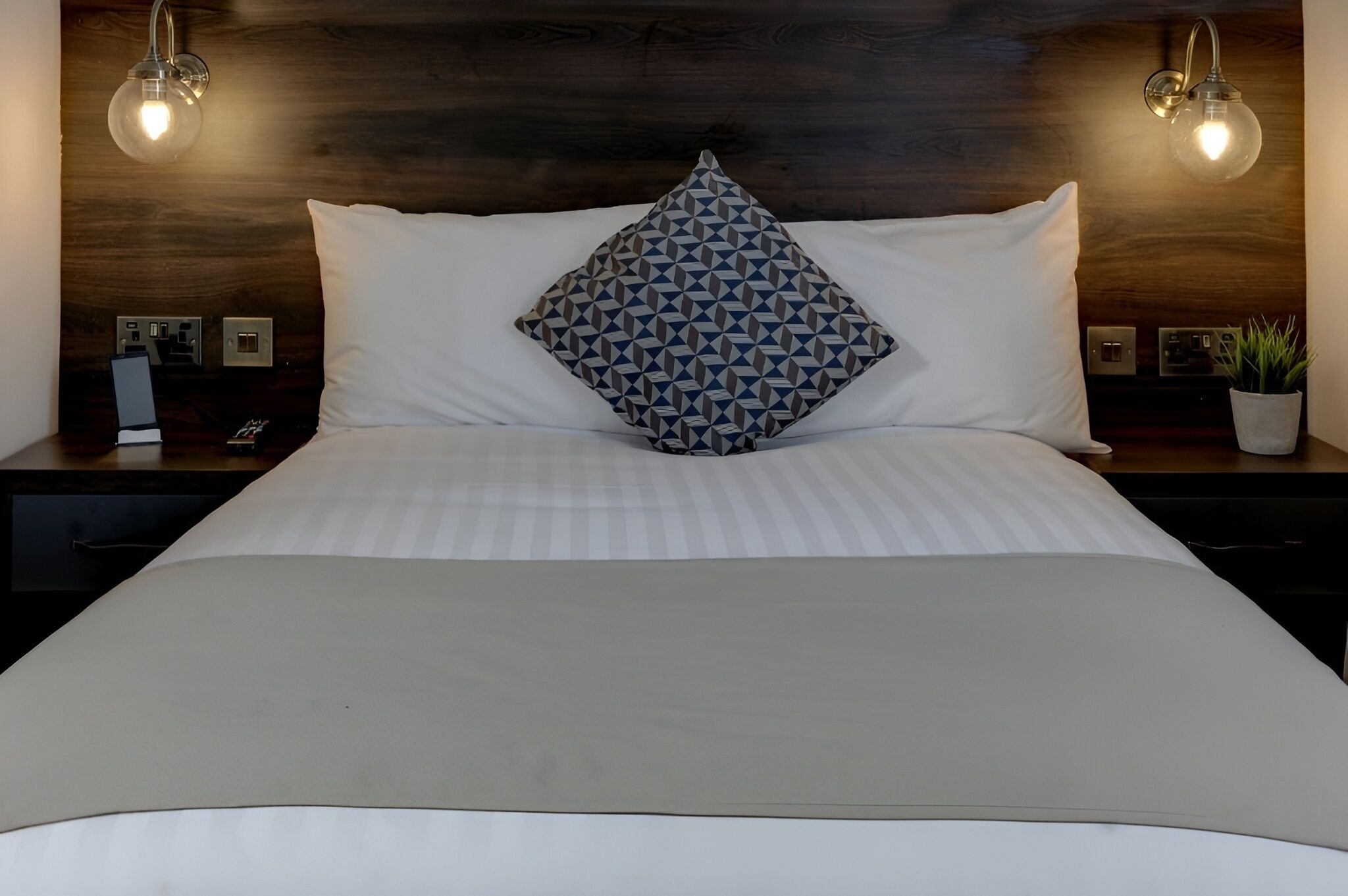 A neatly made bed with a patterned pillow, a wooden headboard, and two wall-mounted lamps.