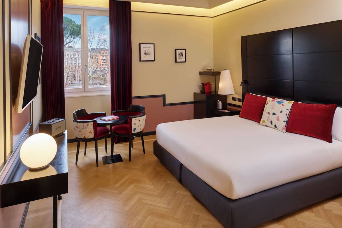 Une chambre d’hôtel avec un grand lit, un bureau avec une lampe, une chaise, une télévision et une fenêtre avec des rideaux.