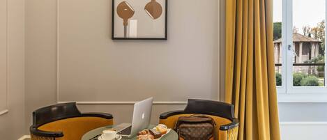Una habitación con dos sillas, una mesa con una computadora portátil, una taza y pasteles. Hay una cita en un marco en la pared y una ventana con cortinas visible.