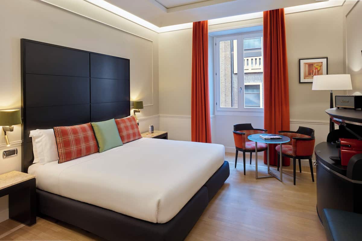 Une chambre d’hôtel avec un grand lit, un bureau avec une chaise, une petite table avec deux chaises, une télévision et une lampe.