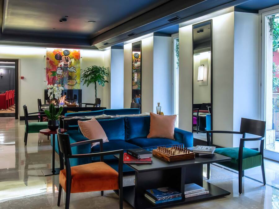Una sala de estar moderna con un sofá azul, un reposapiés naranja y un tablero de ajedrez sobre una mesa de centro. Hay una planta, un espejo y una puerta que da a otra habitación.