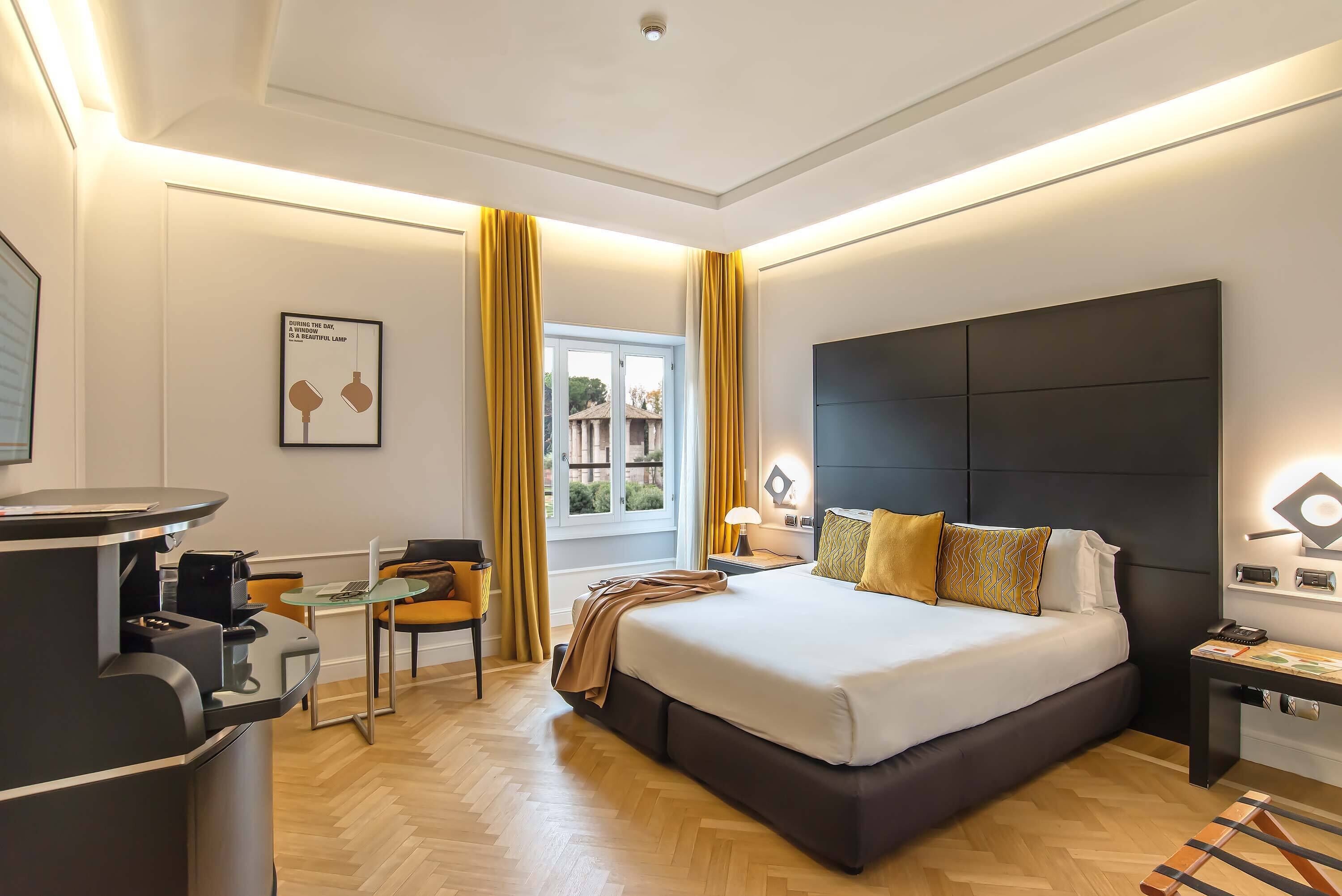 A modern hotel room with a large bed, a desk with a computer, a chair, and a window with curtains.