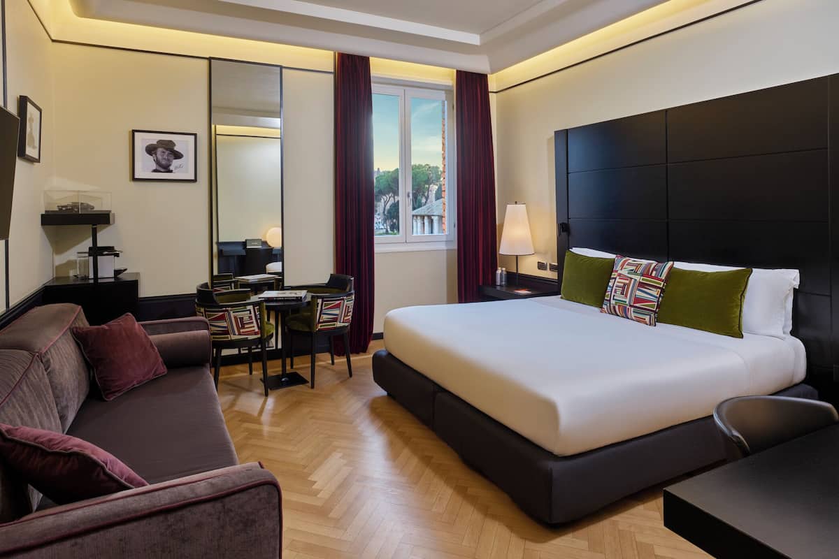 Une chambre d’hôtel moderne avec un grand lit, un canapé, un bureau et une chaise. On y trouve une fenêtre avec des rideaux, un miroir et une lampe.