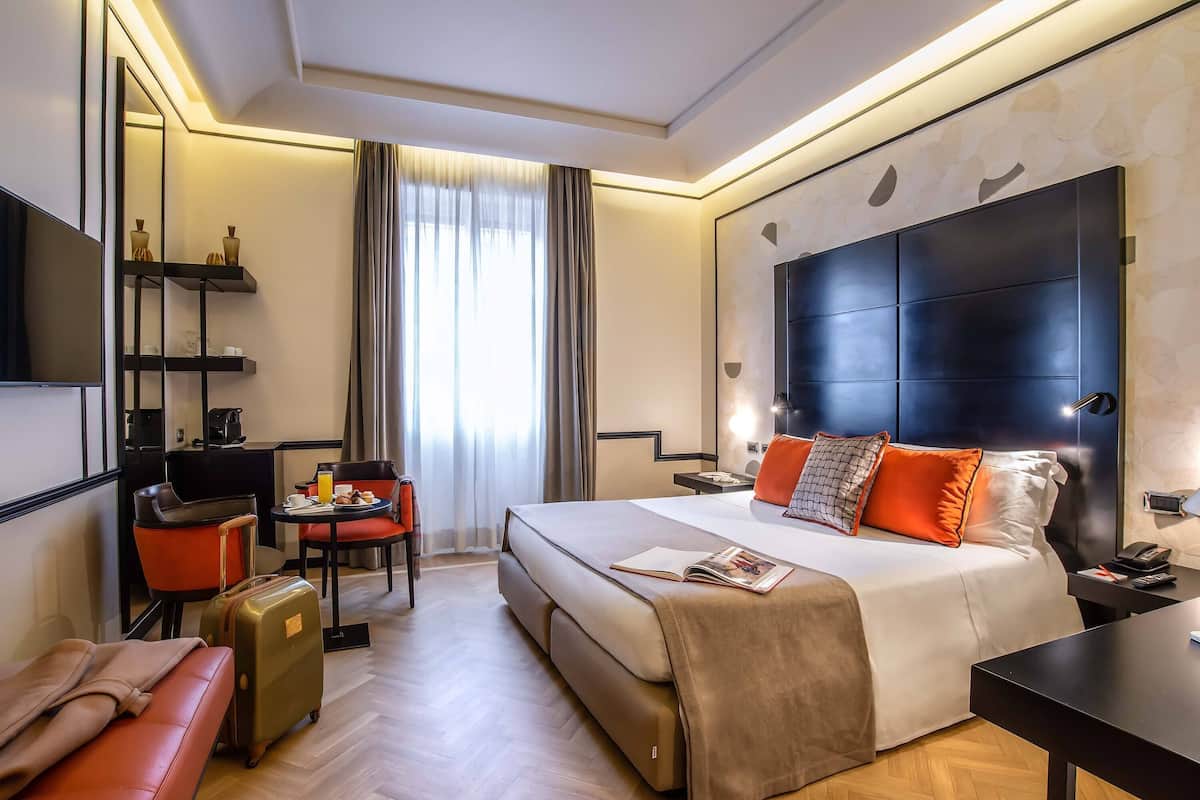 A hotel room with a bed, bedside table, lamp, chair, desk, and a suitcase.